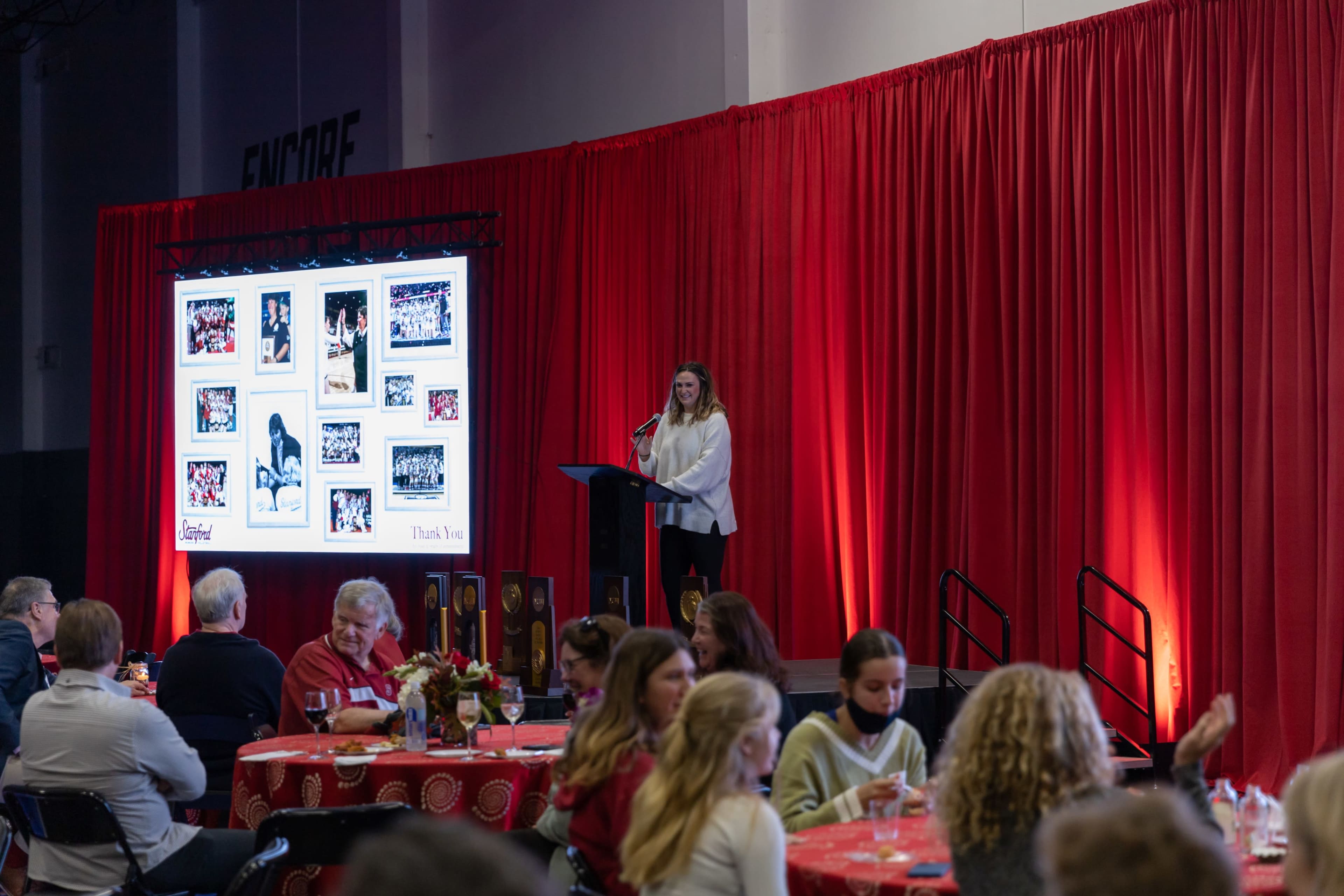 A woman stands at a podium speaking to an audience during an event, with a large screen displaying photos in the background.