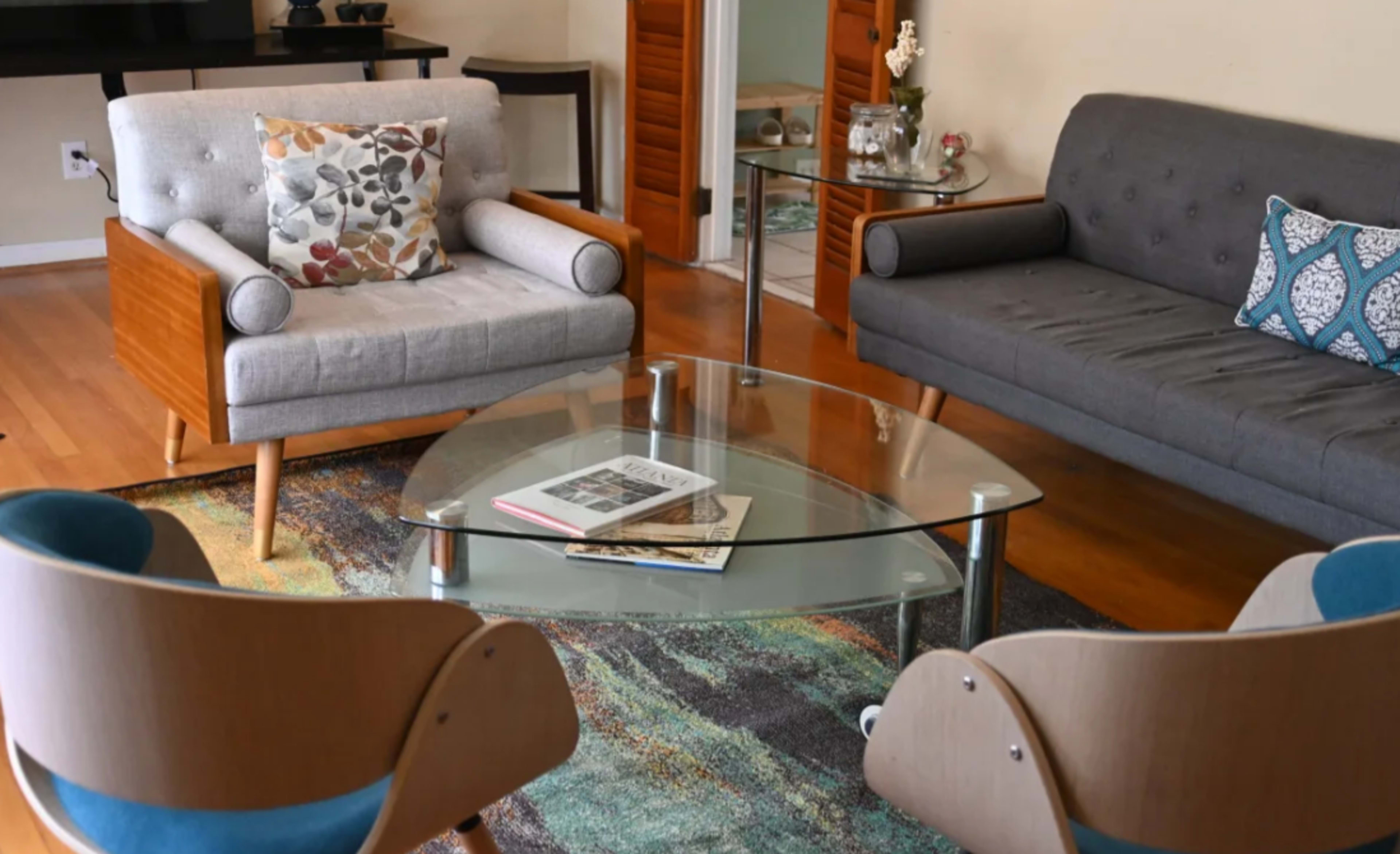 A living room features a glass coffee table surrounded by two modern chairs and two sofas, with a decorative rug underneath.
