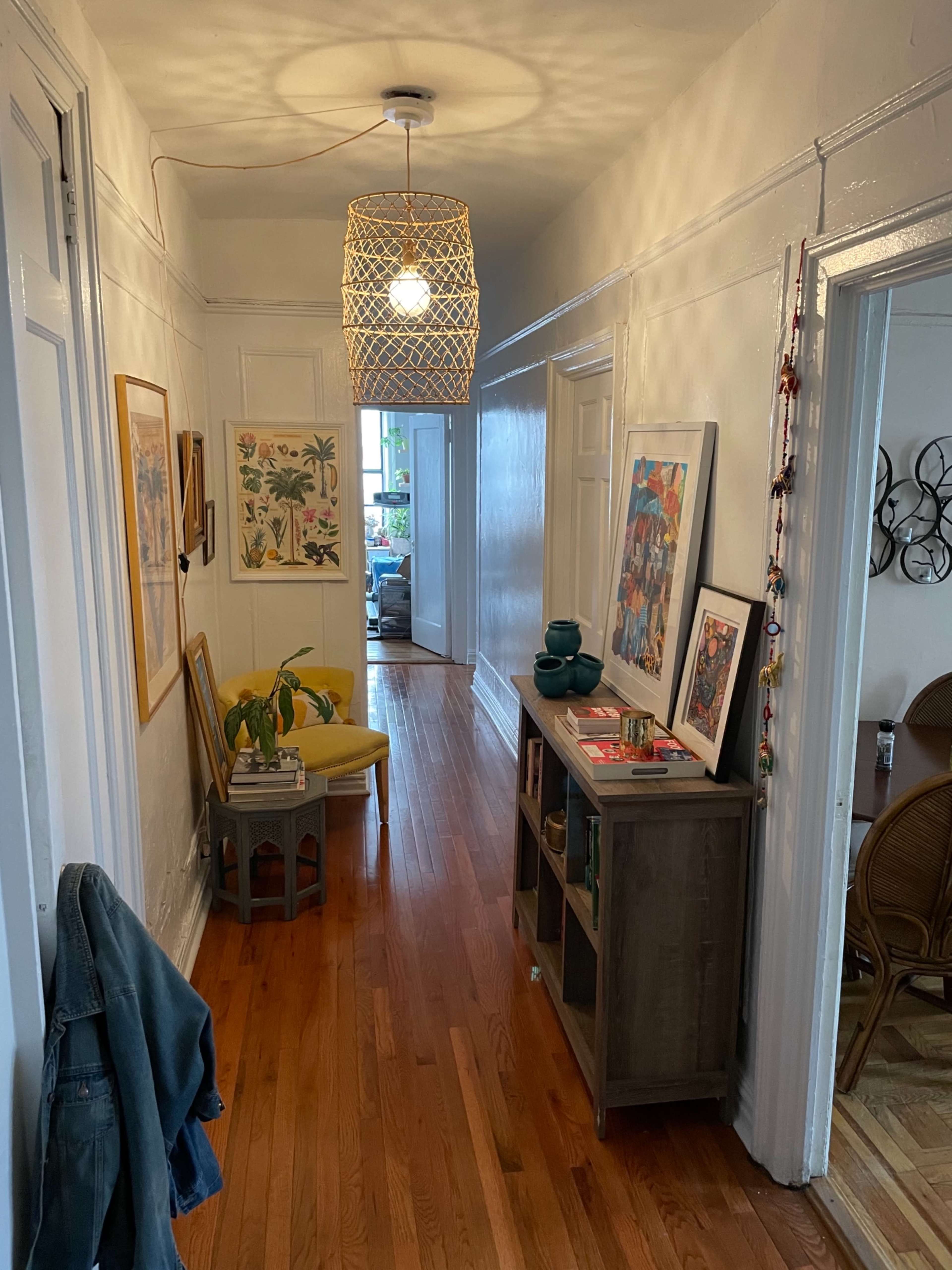 A narrow hallway features a woven light fixture, artwork on the walls, a small round table with a plant, and a storage unit beside a chair.