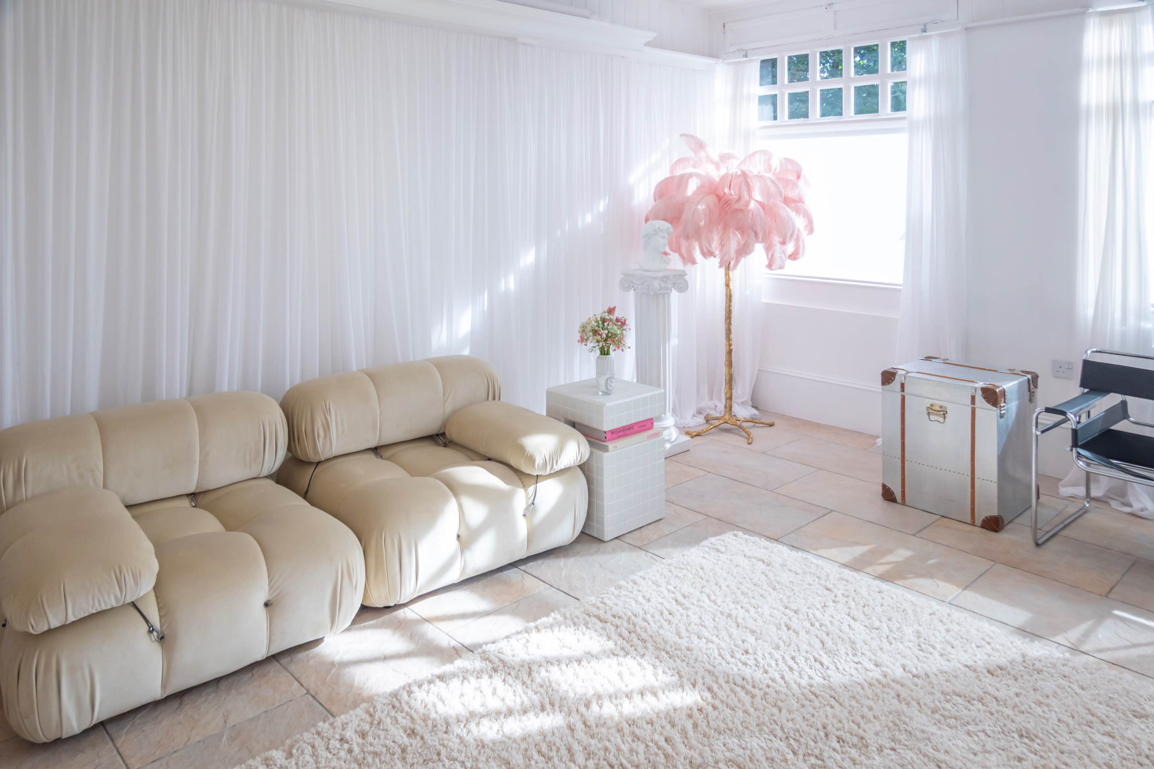 A bright, minimalist living space with a cream-colored sofa, a small table topped with flowers, a pink decorative tree, and a metallic trunk.