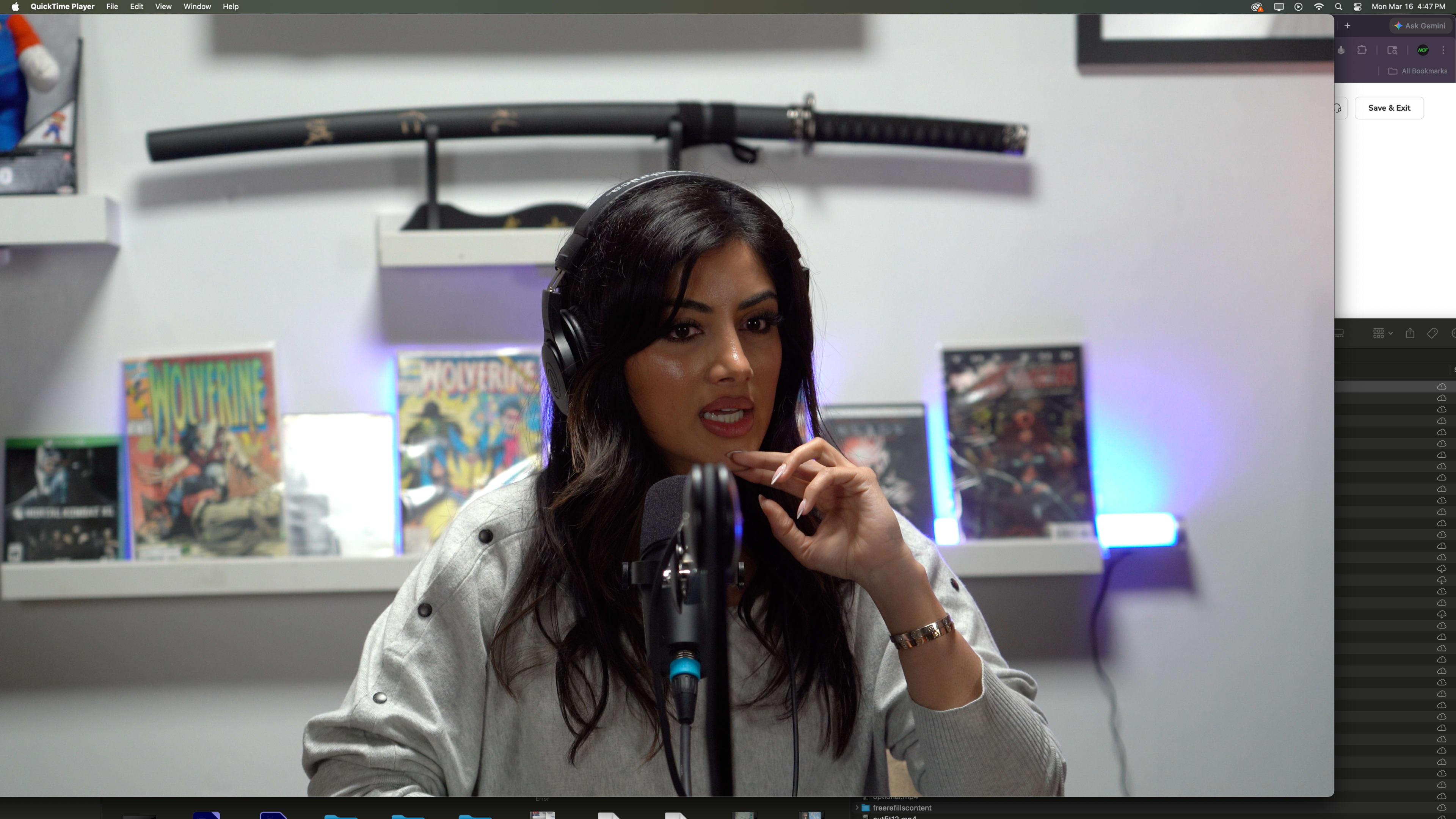 A woman is speaking into a microphone while wearing headphones, with comic book covers displayed on a shelf behind her.