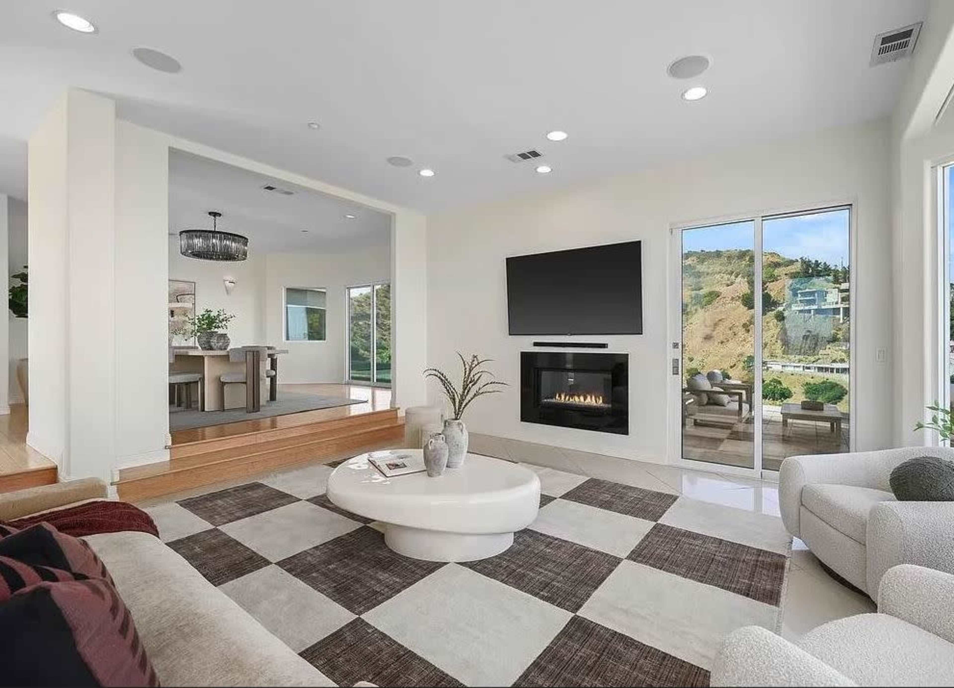 A living room features a round coffee table on a patterned rug, a fireplace, and large windows that open to a view of the outdoors.
