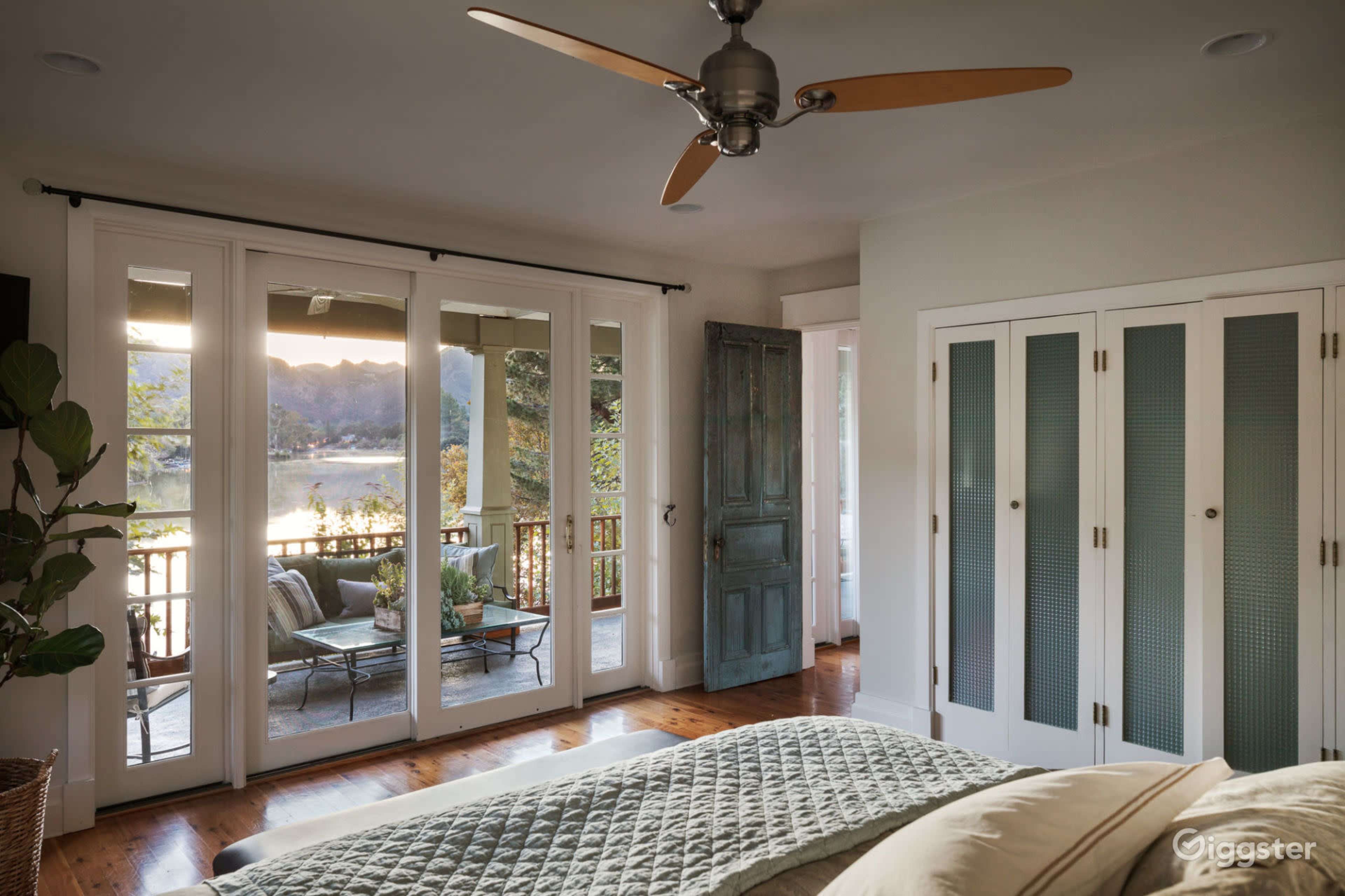 A cozy bedroom features a large glass door leading to a balcony with views of a lake, complemented by a ceiling fan and a wooden door.