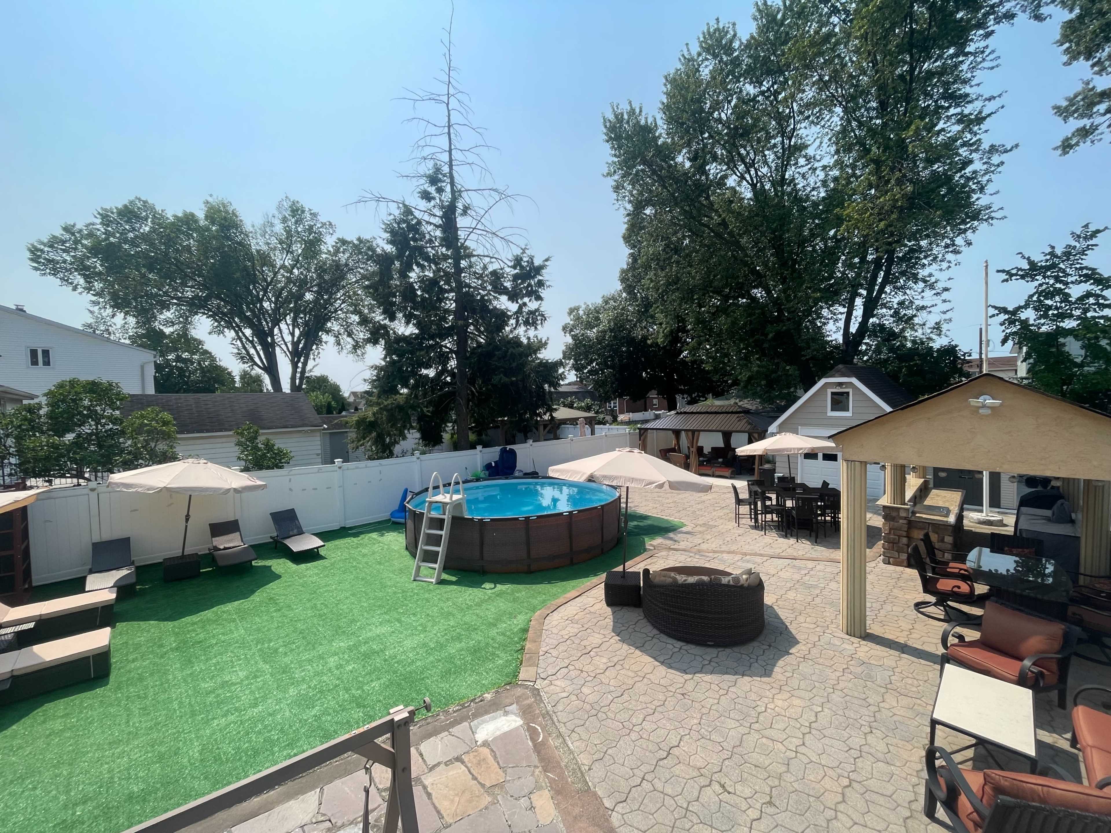 The image shows a backyard featuring a circular above-ground pool surrounded by lounge chairs, umbrellas, and a patio area with seating.