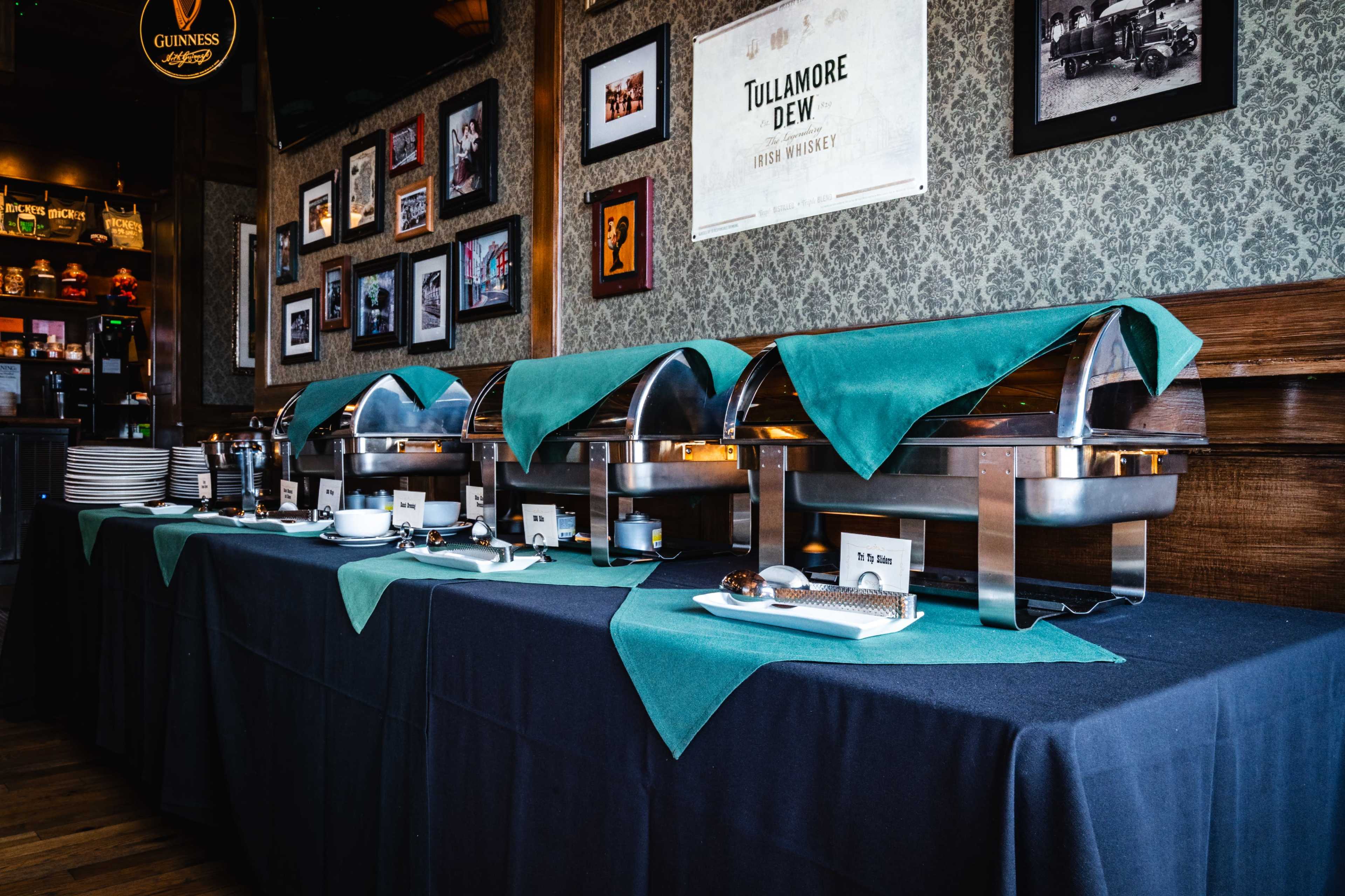 The image shows a buffet table set with metal chafing dishes covered by green cloths, accompanied by plates and decorative elements against a patterned wall.