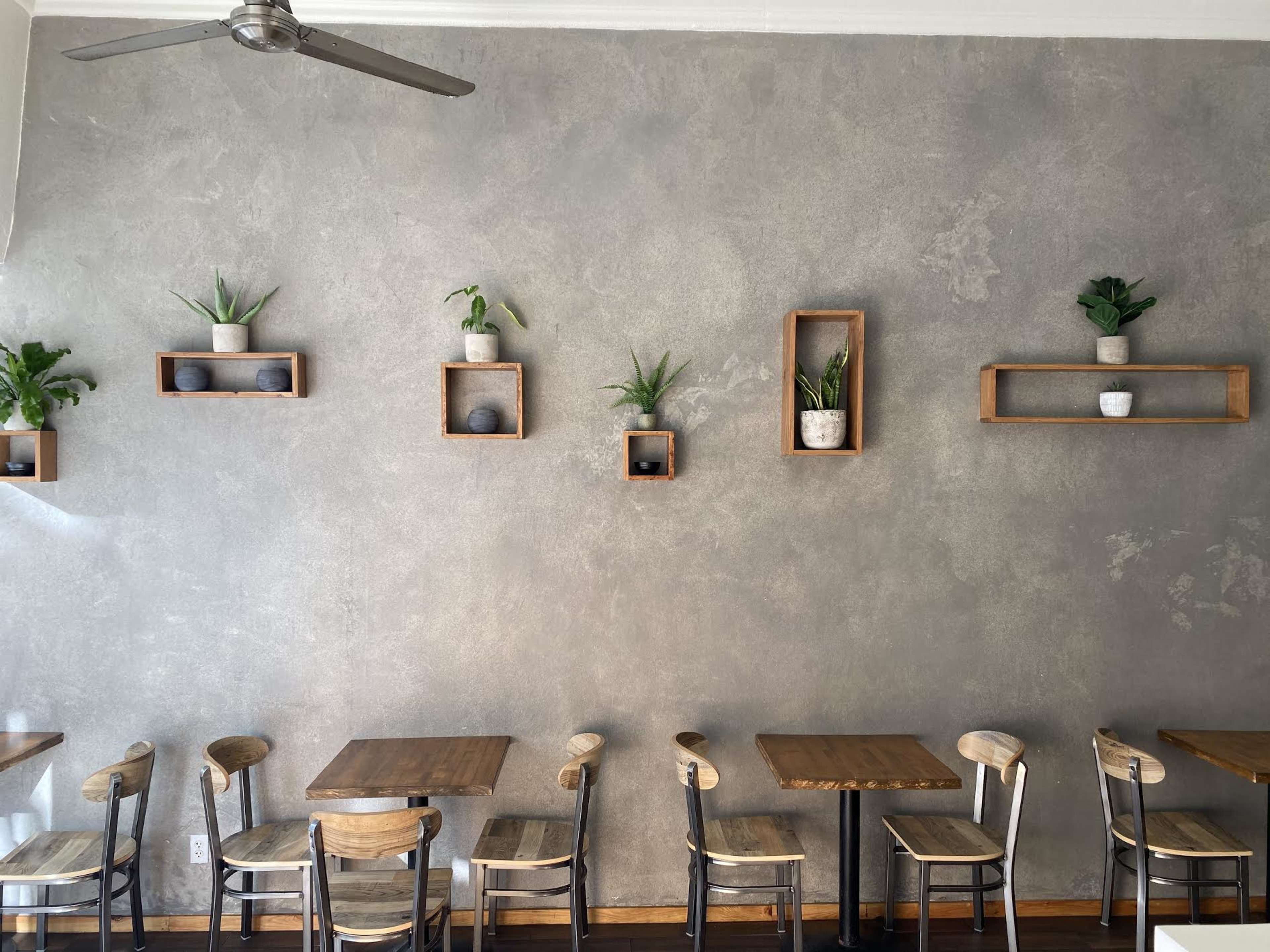 A café interior featuring wooden tables and chairs, with a textured gray wall adorned with floating shelves displaying potted plants.
