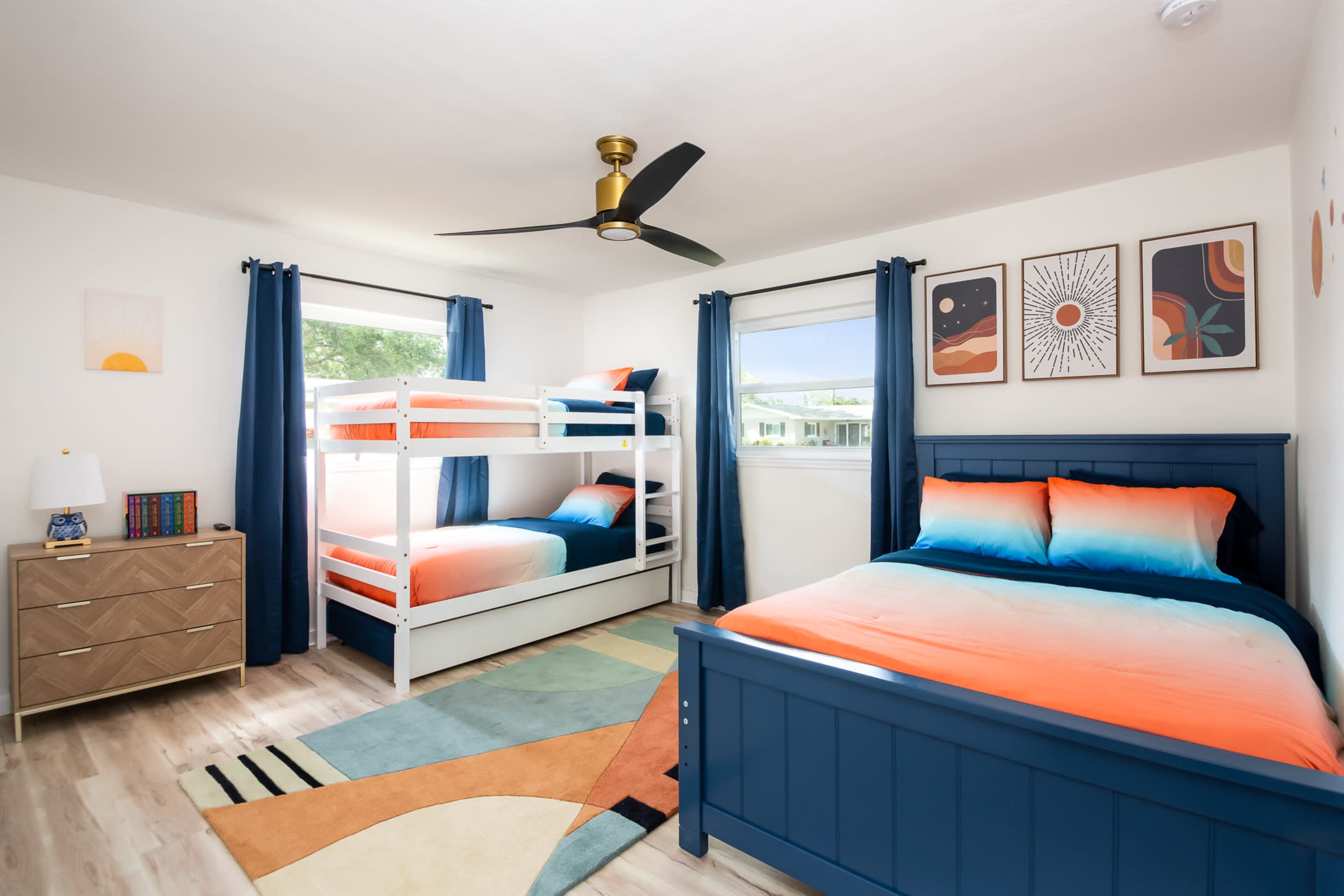 The room features a bunk bed and a single bed with orange and blue bedding, paired with a dresser and decorative wall art.