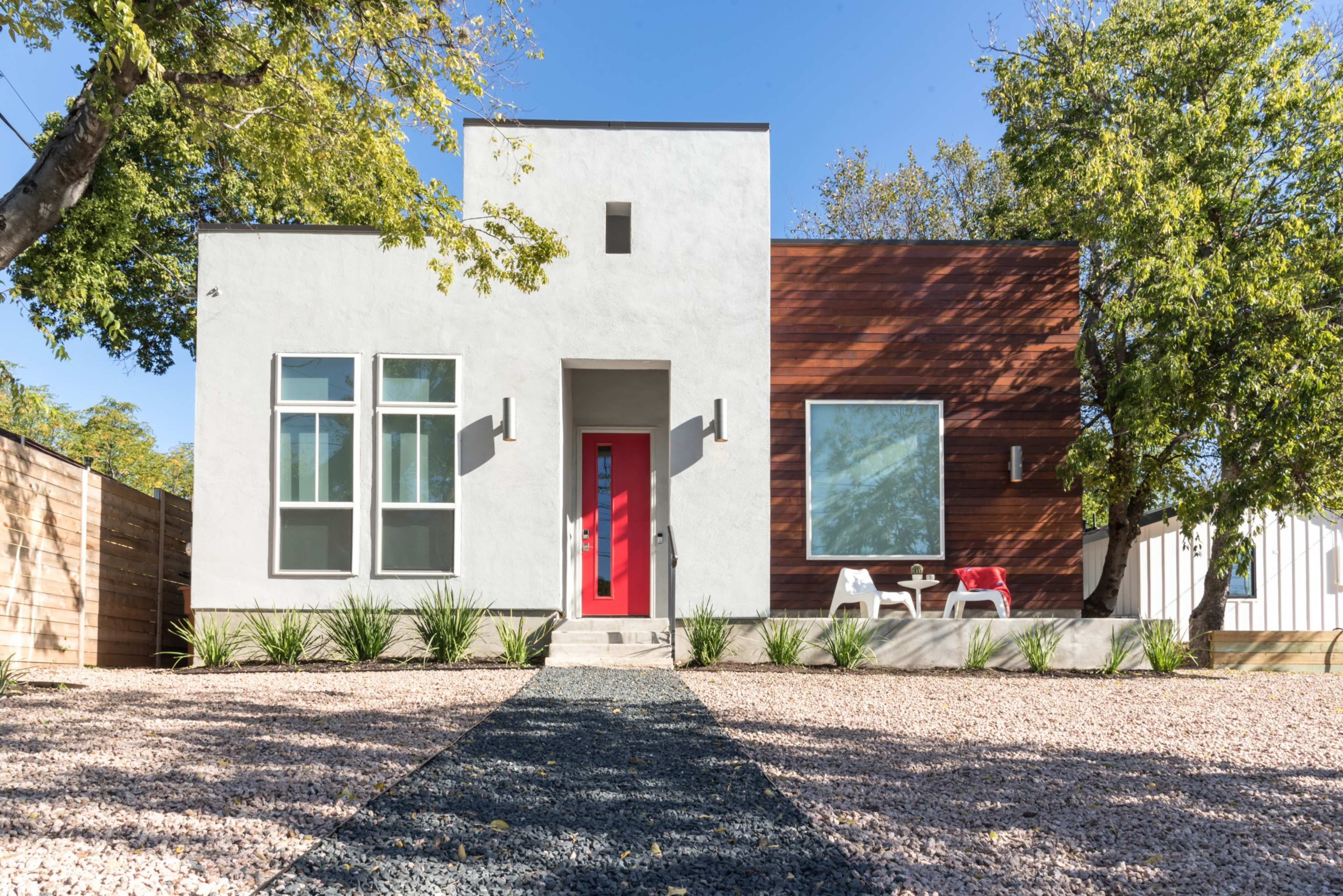 A modern house features a white exterior with wooden accents, a bright red door, and landscaped gravel pathways.