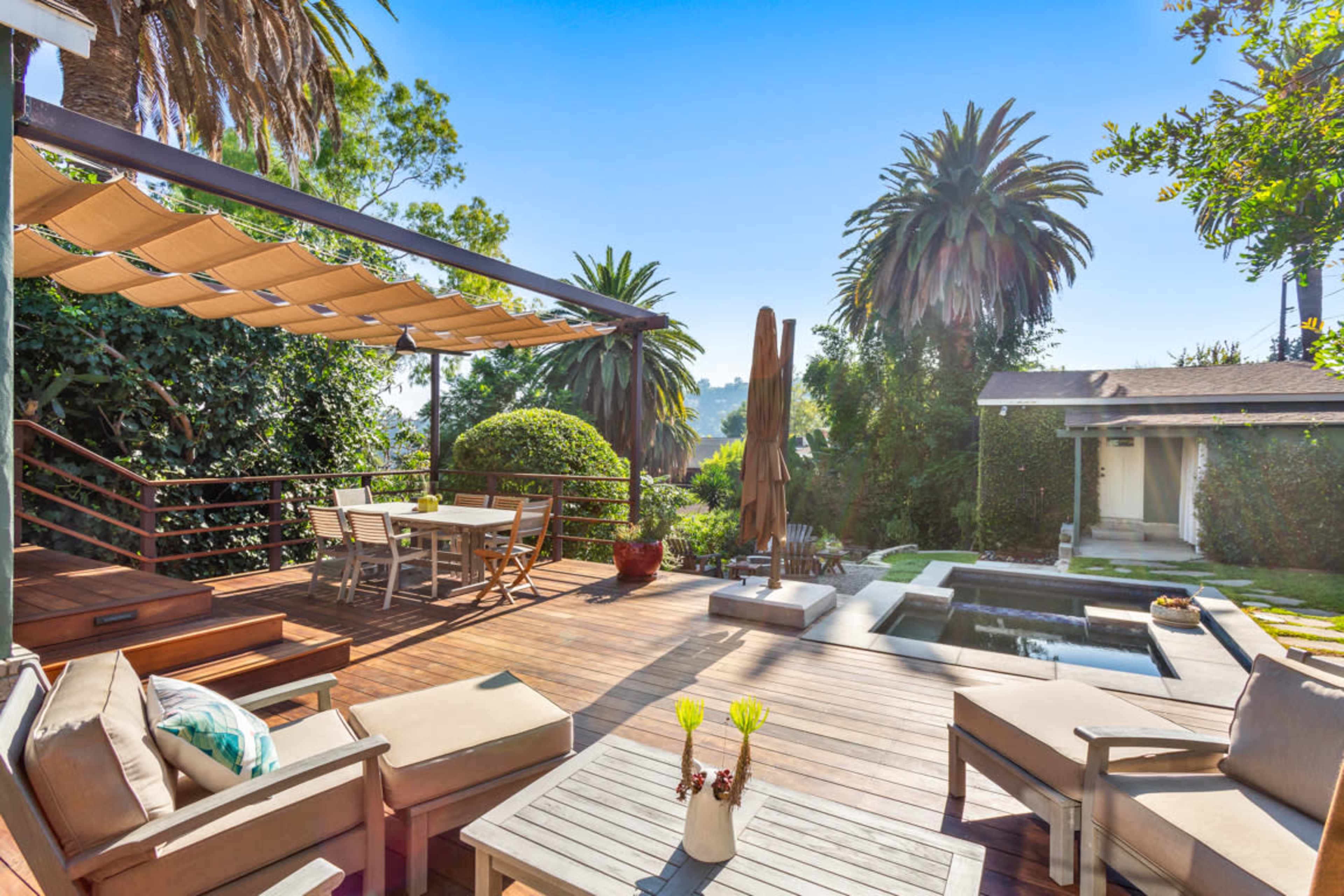 The image shows a landscaped outdoor patio area with a wooden deck, seating, a dining table, and a small pool surrounded by palm trees.