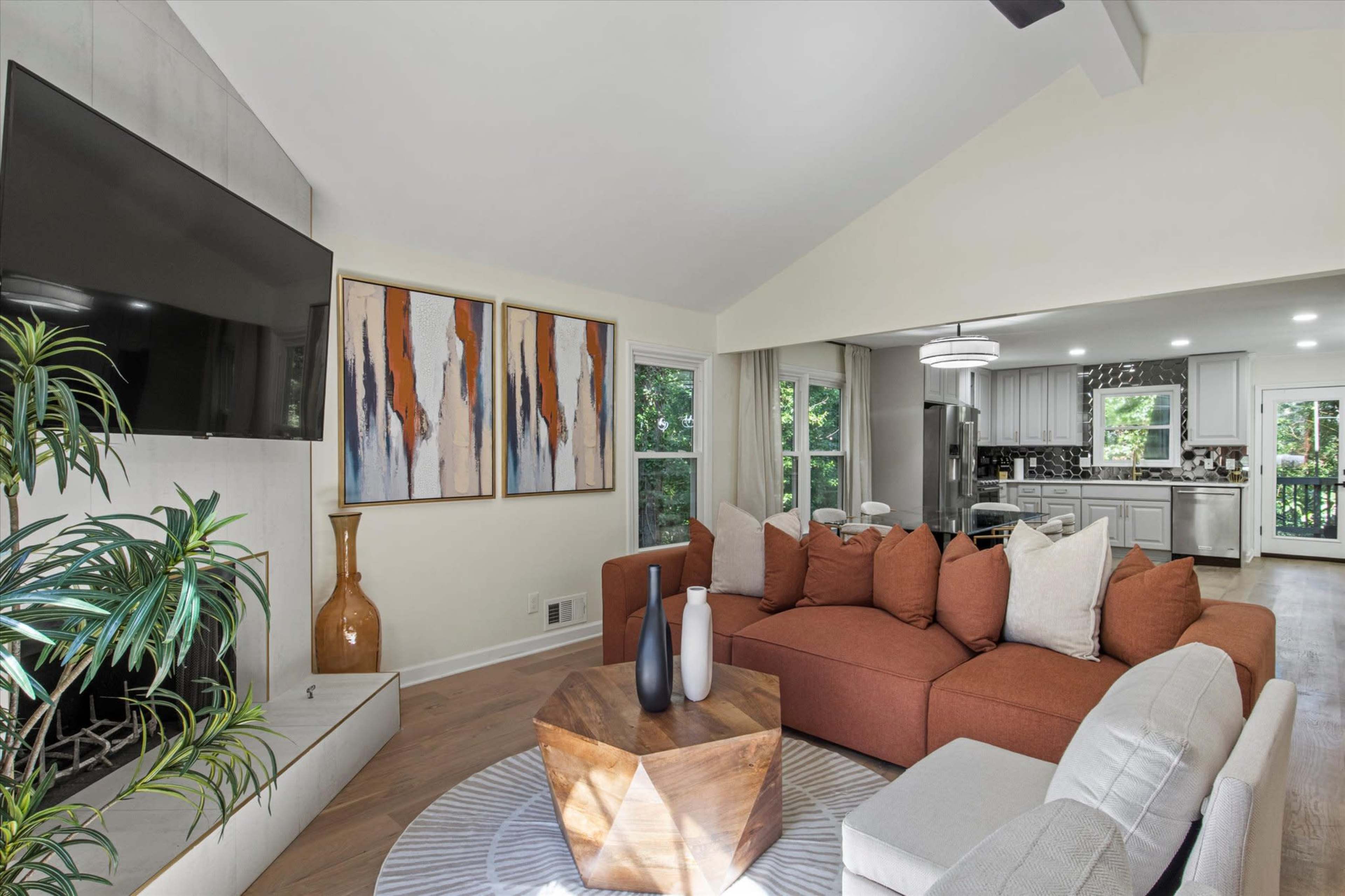 The image shows a modern living room with a large sectional sofa, an artistic wall decor above a fireplace, and an open kitchen in the background.