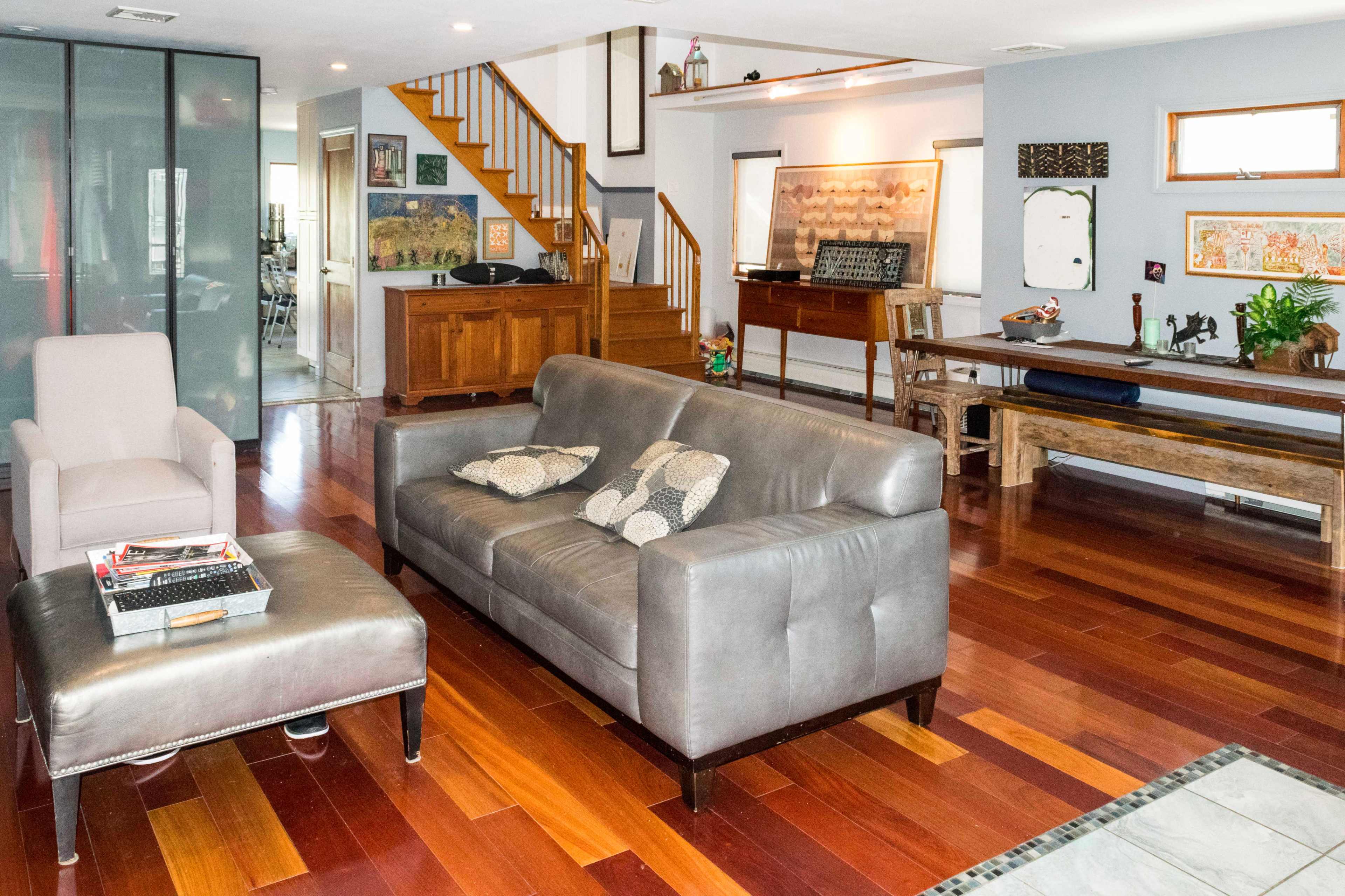 The living area features a gray leather sofa and ottoman, wooden stairs leading to an upper level, and various decorative items displayed on the walls and furniture.