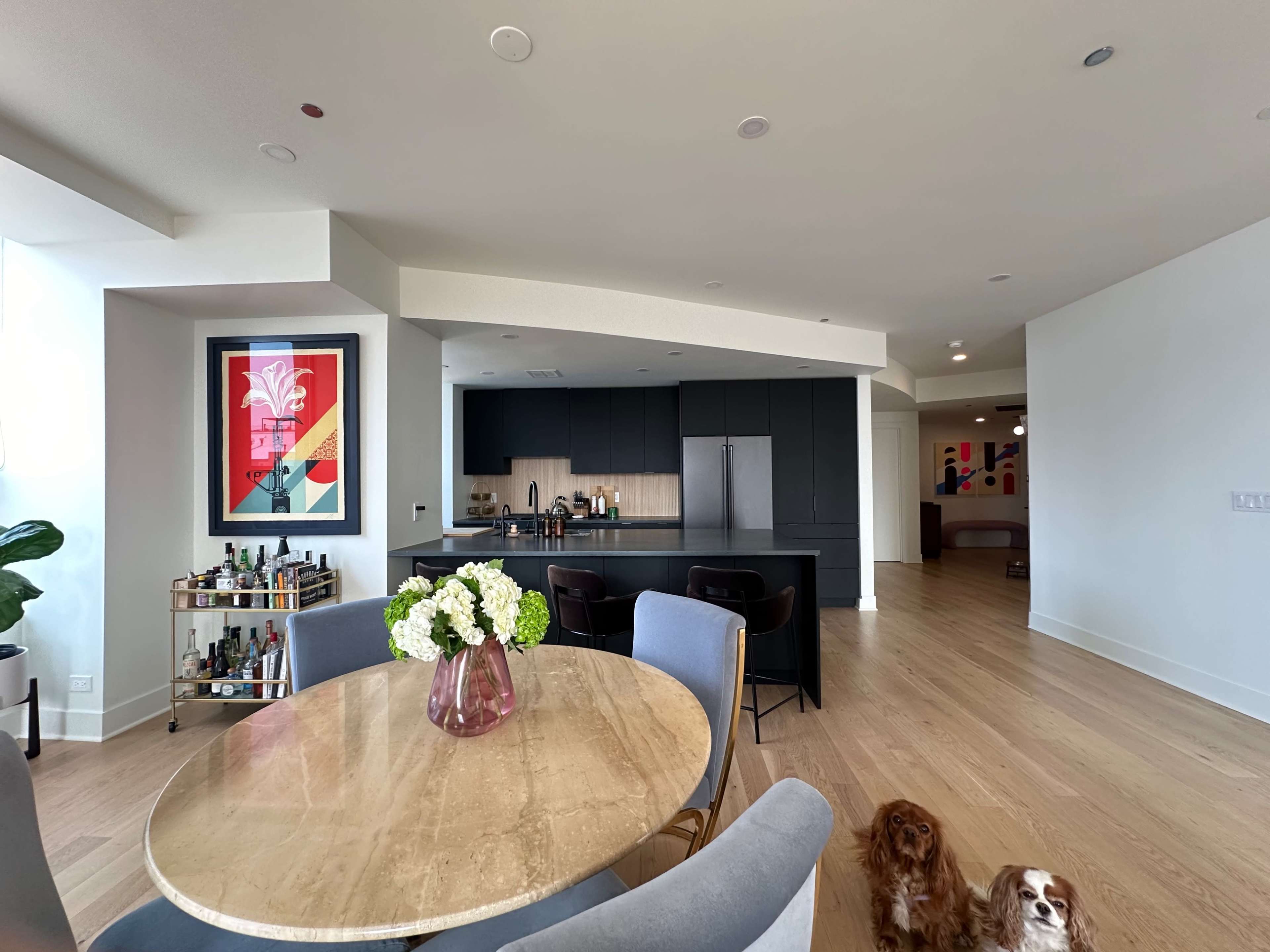 A modern kitchen and dining area feature a round table with a floral arrangement, a bar cart, and two dogs on the floor.