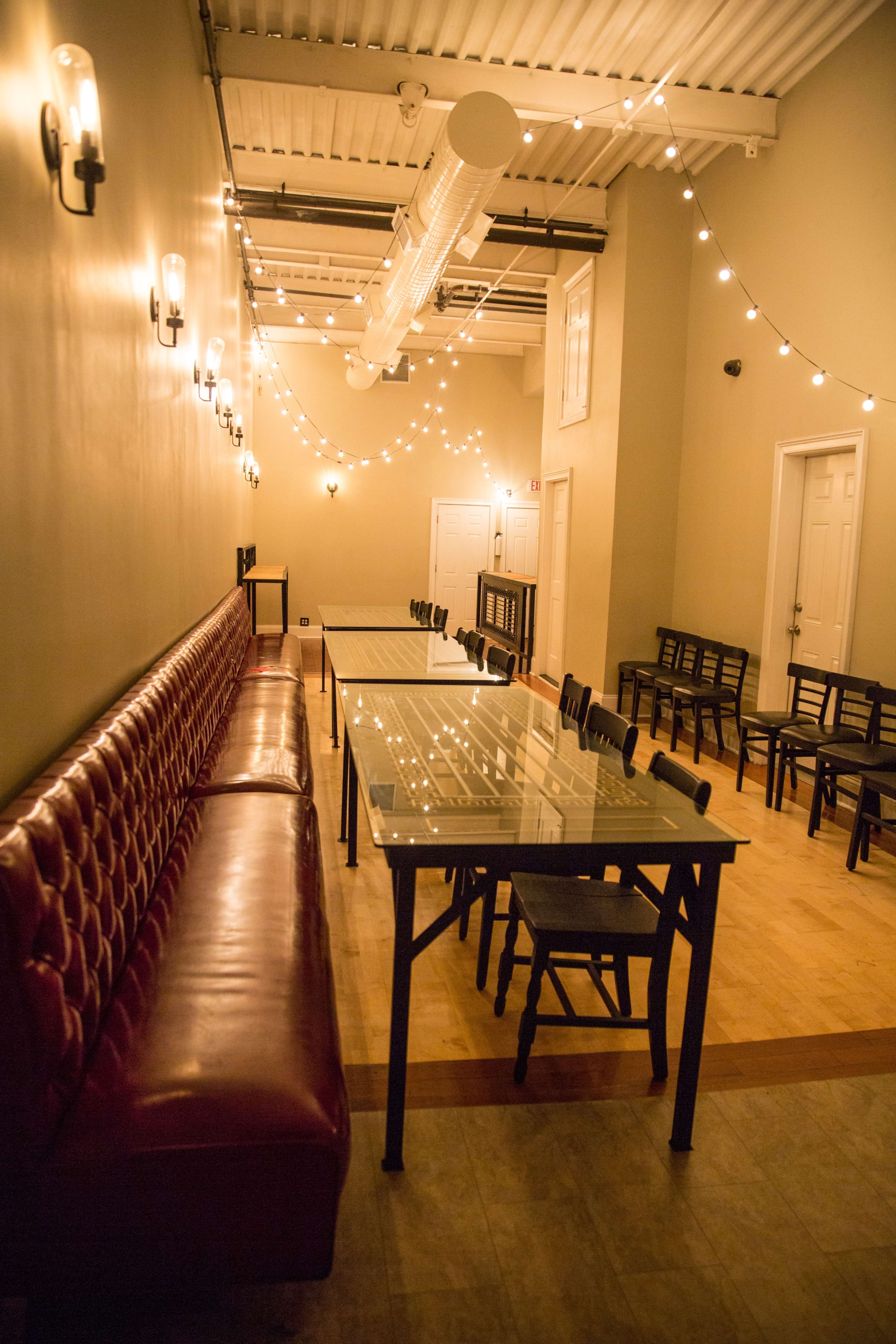 The image shows a long, narrow room with a red cushioned bench along one wall, glass-topped tables arranged in the center, and wooden chairs lined up against the opposite wall, illuminated by string lights overhead.