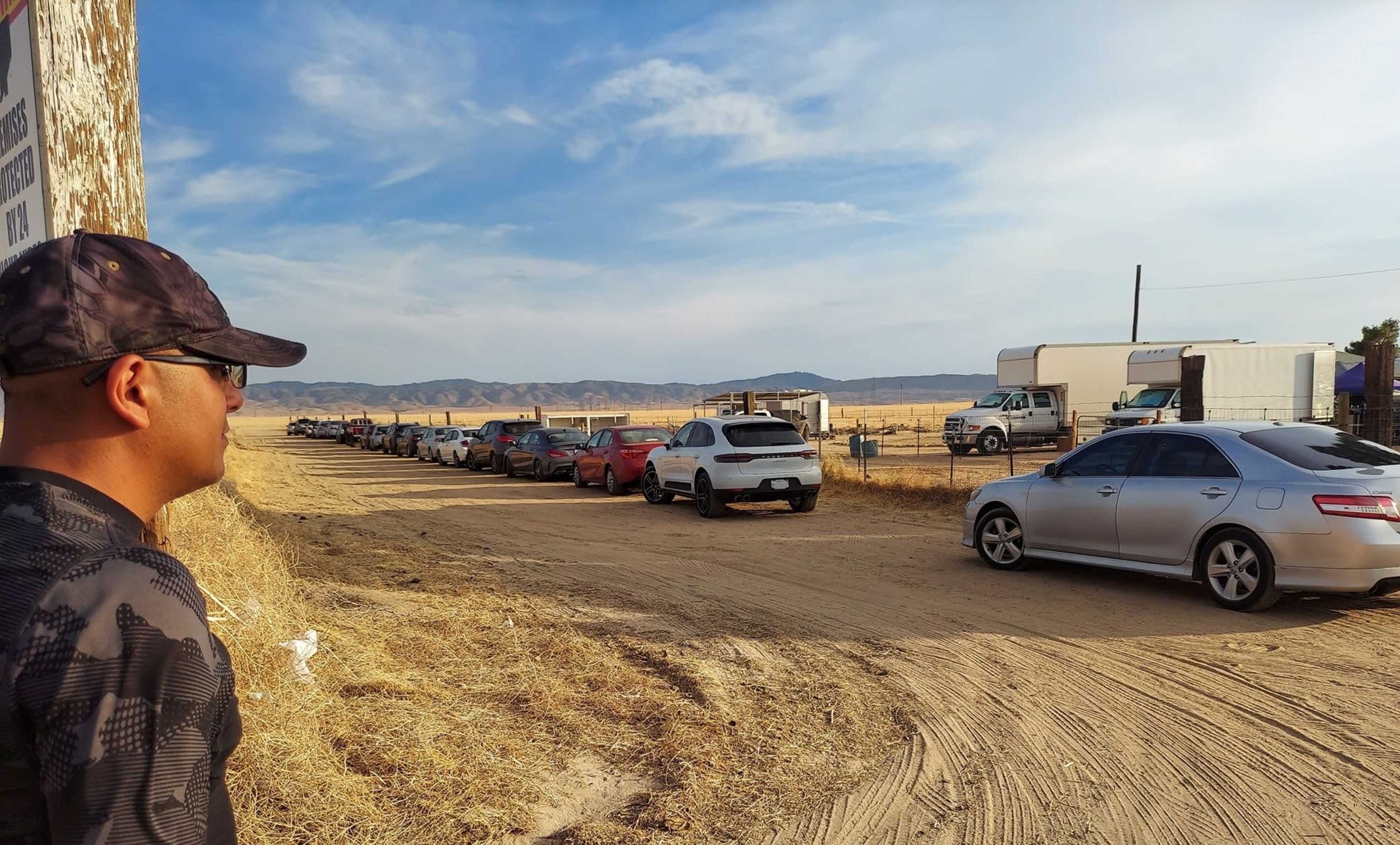 A man stands on a dirt road overlooking a line of parked cars against a backdrop of open fields and distant mountains.