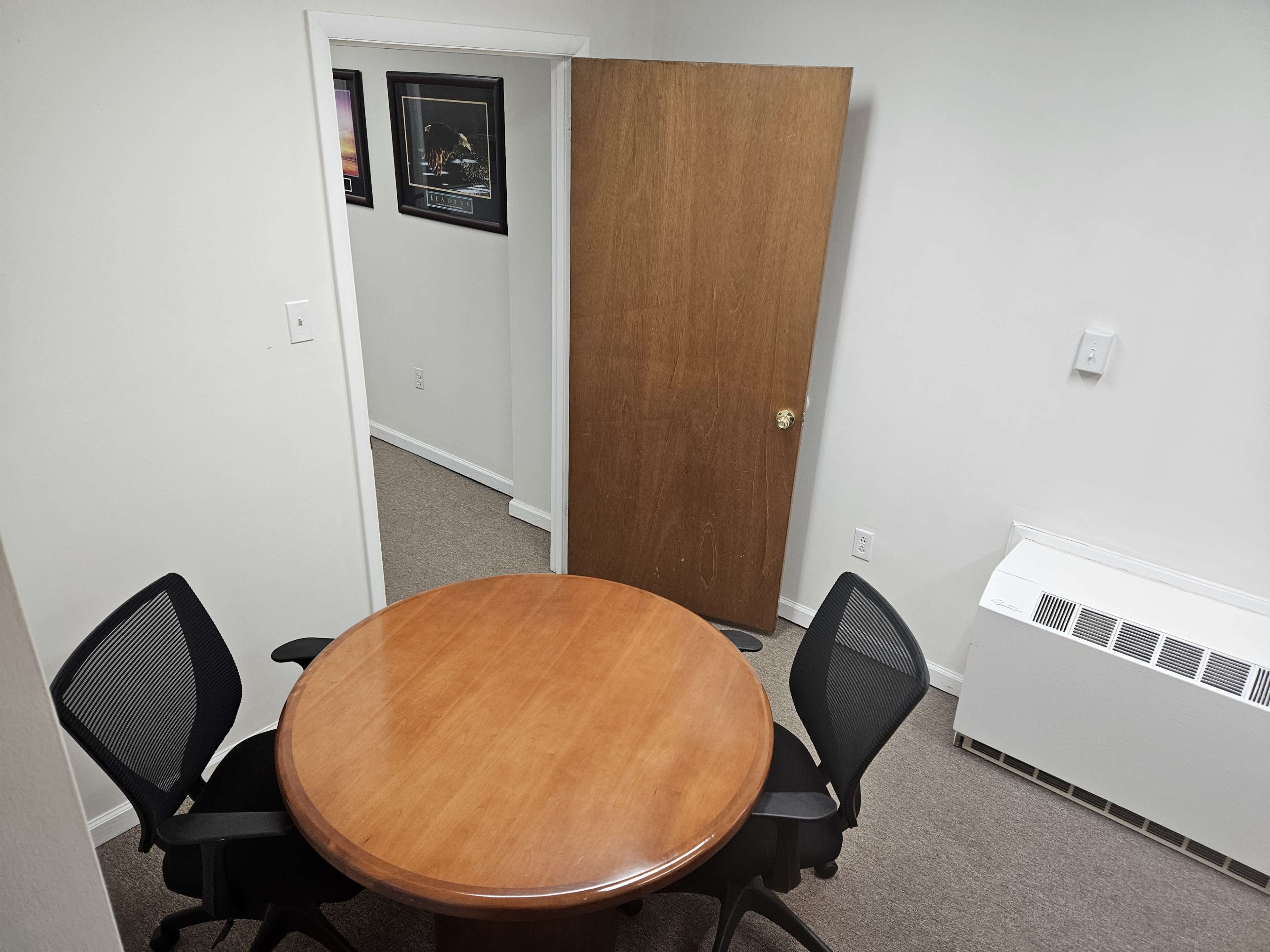 The image shows a small meeting room featuring a round wooden table with two black chairs and a partially open door leading to another room.