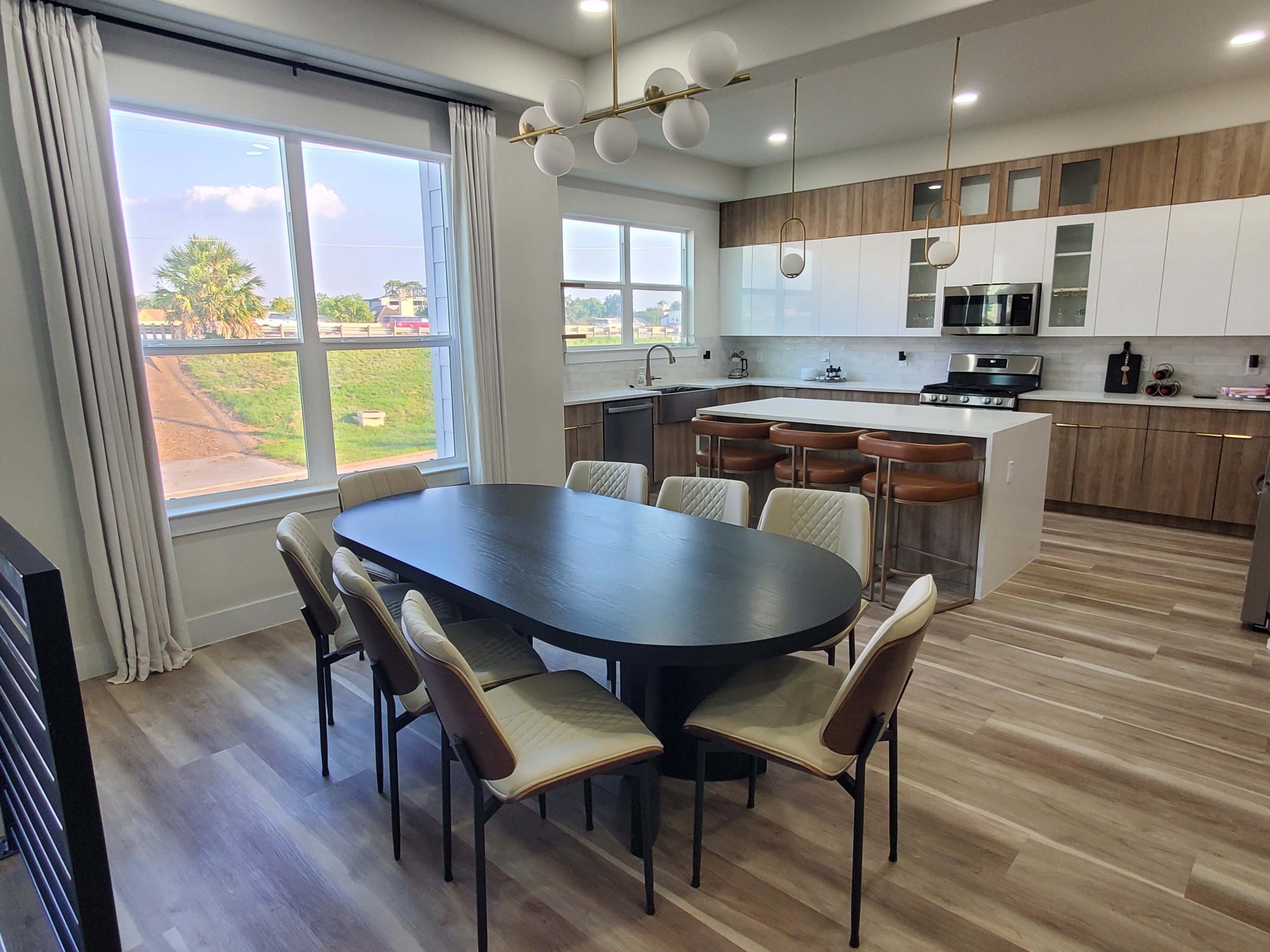 The image shows a modern kitchen and dining area featuring a large oval table surrounded by chairs, with a view of a landscape through a window.
