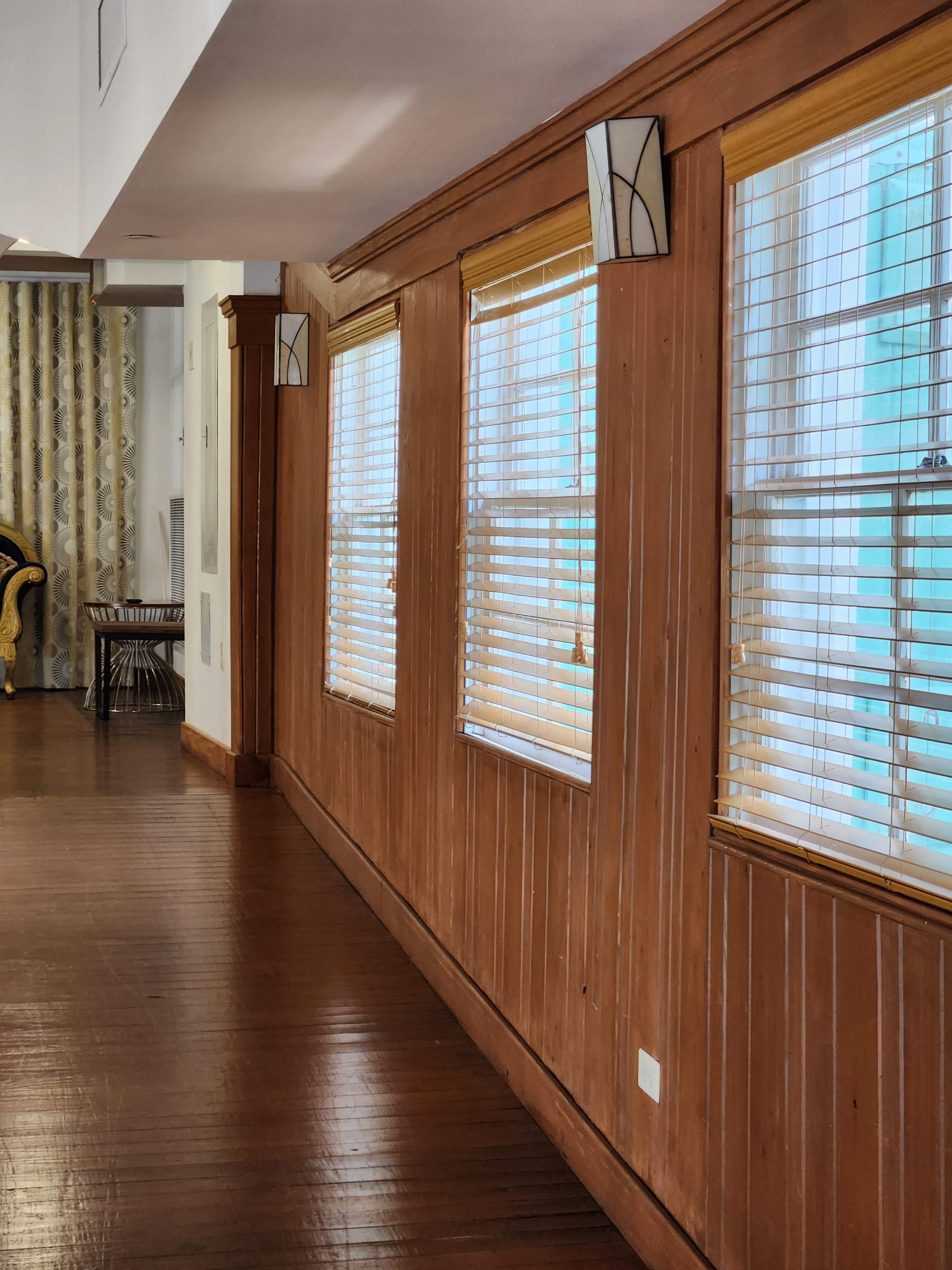A wooden-paneled wall with three windows featuring blinds, alongside a curtain and a glimpse of furniture in a well-lit interior space.