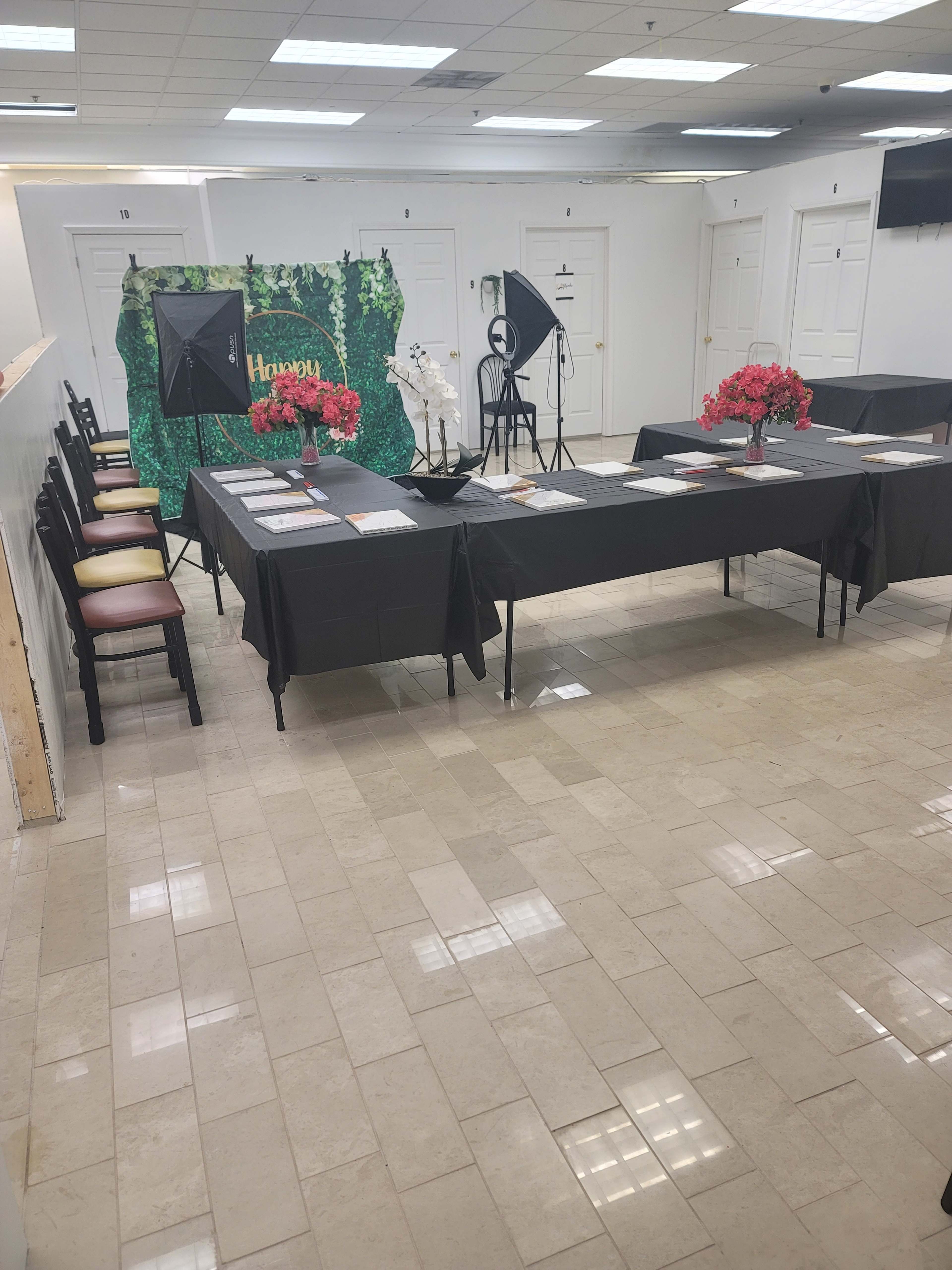 A spacious room features a long table covered with documents, chairs aligned on one side, and a decorative backdrop with greenery and flowers on the opposite wall.