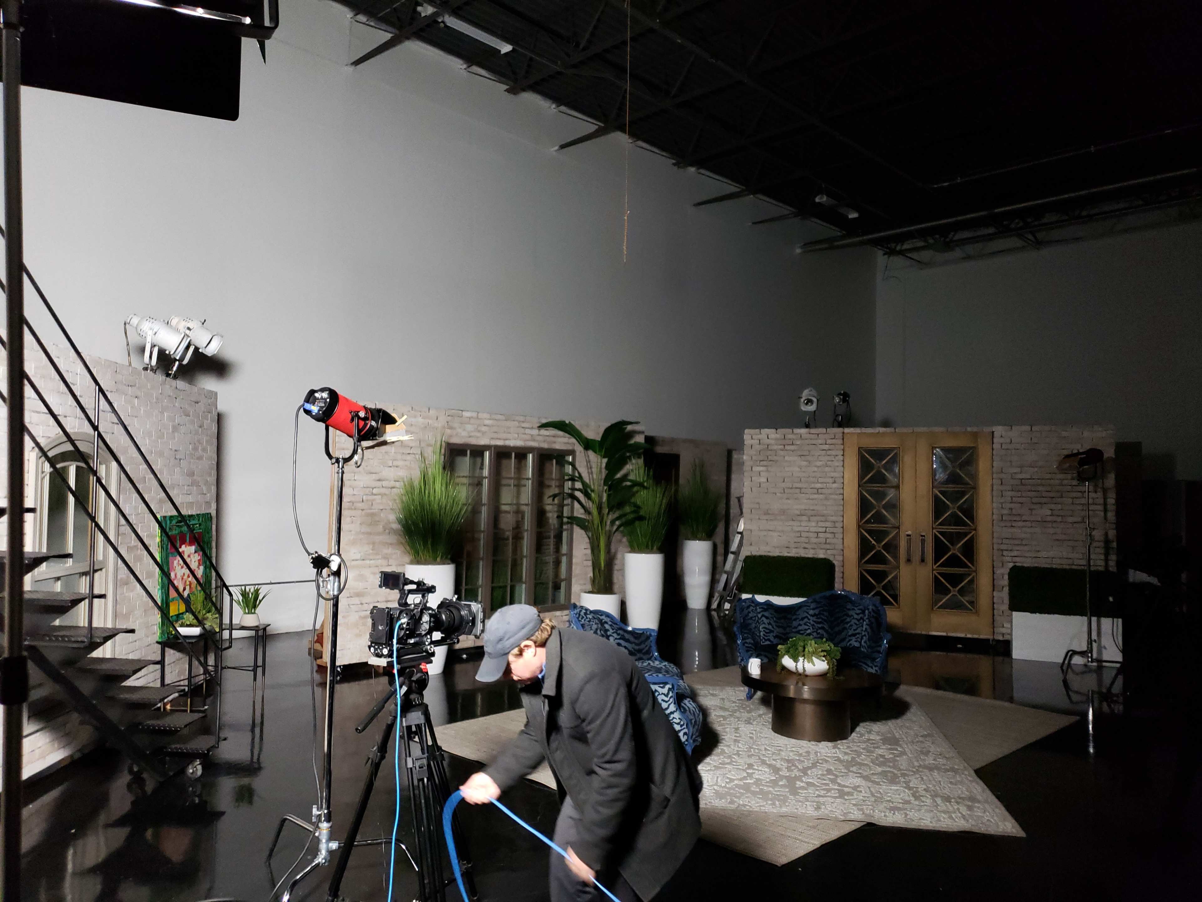 A film crew sets up equipment in a studio featuring a modern living space with large windows, a patterned rug, and various indoor plants.