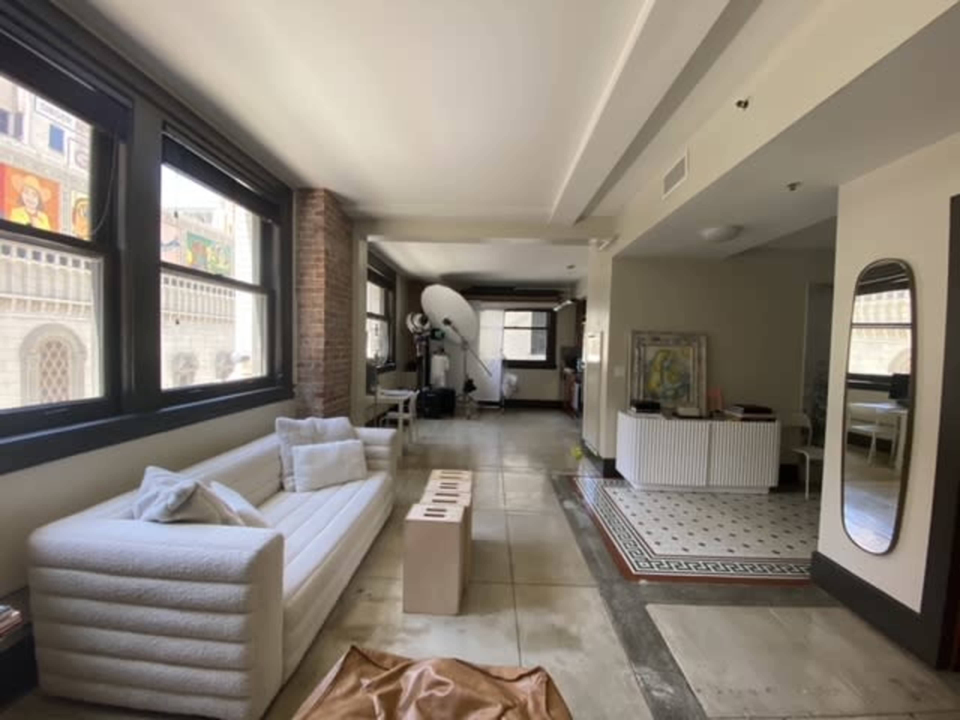 The image shows a spacious, modern loft with large windows, a white sofa, minimalist decor, and a mix of tiled and concrete flooring.