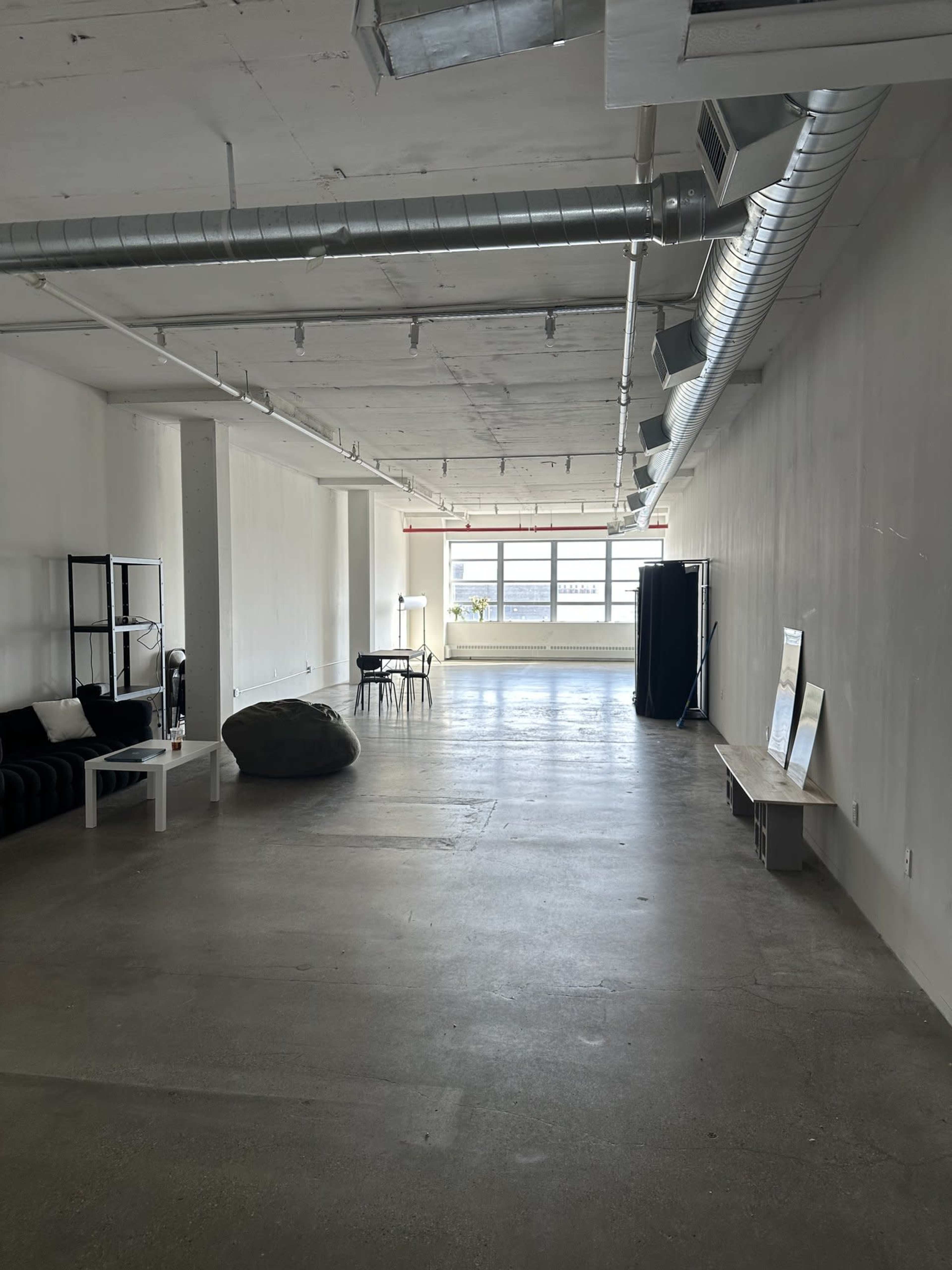 The image shows an empty, light-filled room with a concrete floor, large windows, and minimal furniture including a sofa, a chair, and a bench.