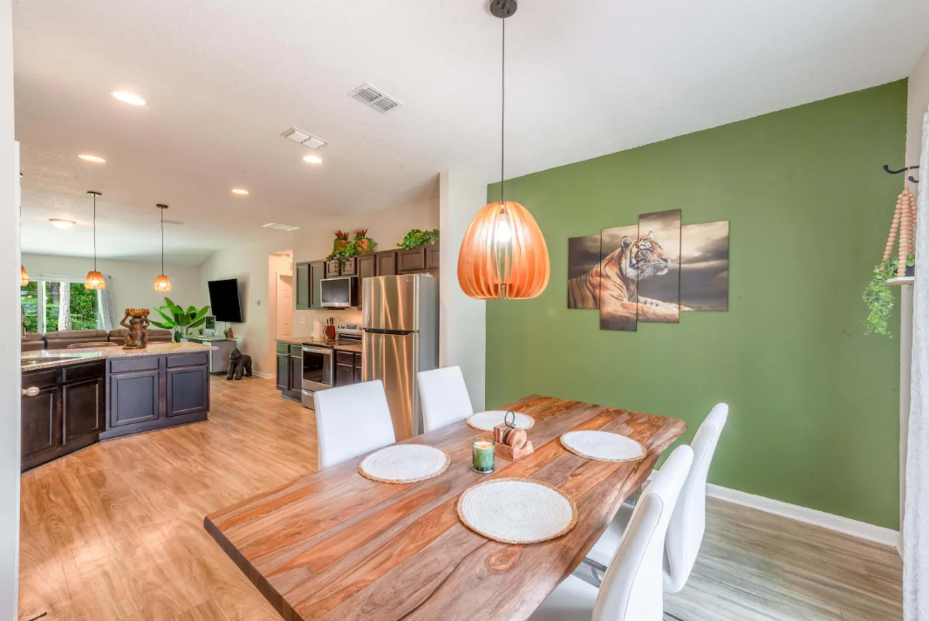 A modern dining area features a wooden table set for four, with a green accent wall and a framed tiger artwork.