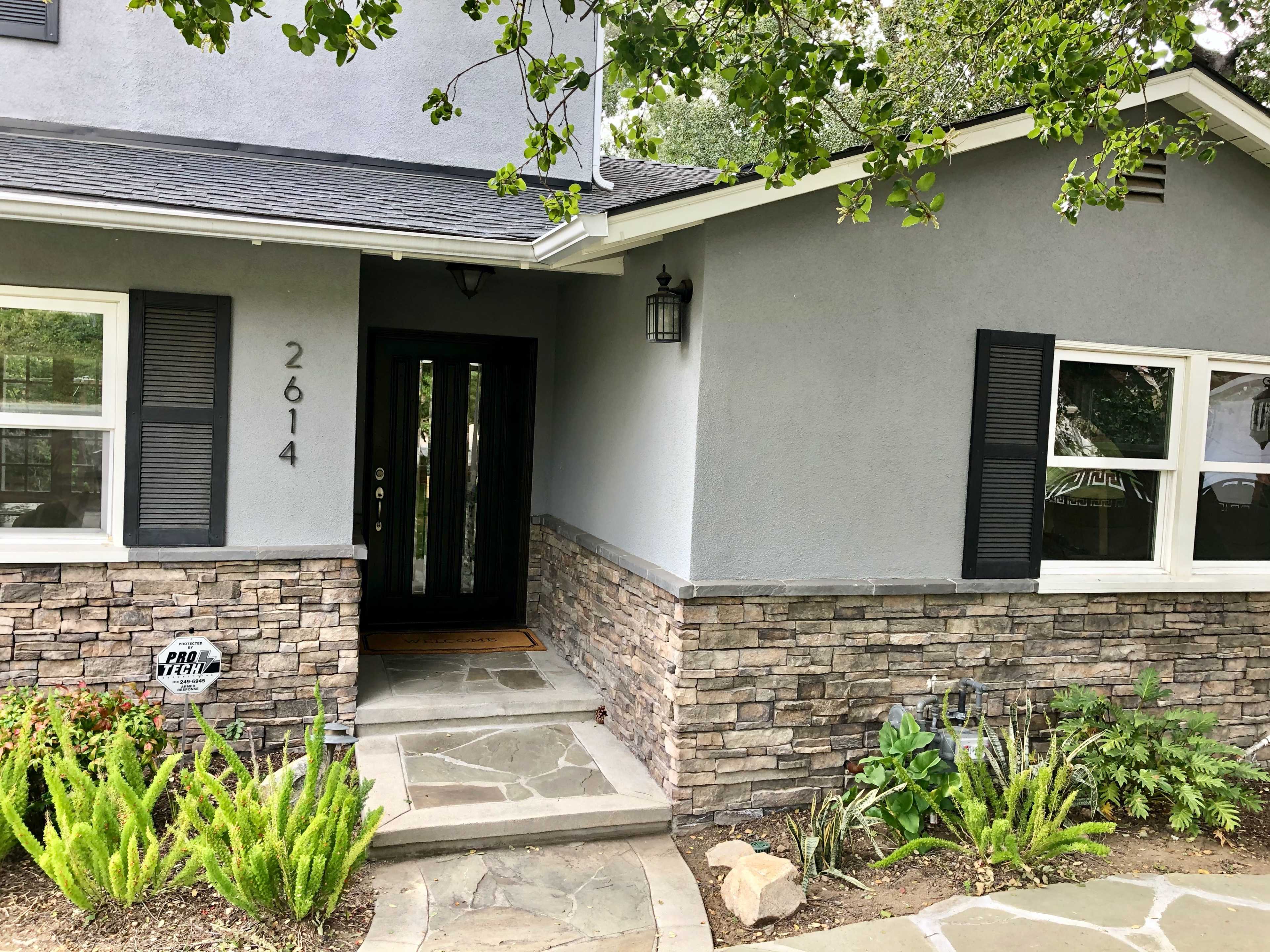 The image shows a home entrance with a gray facade, stone accents, and a landscaped pathway leading to the front door.