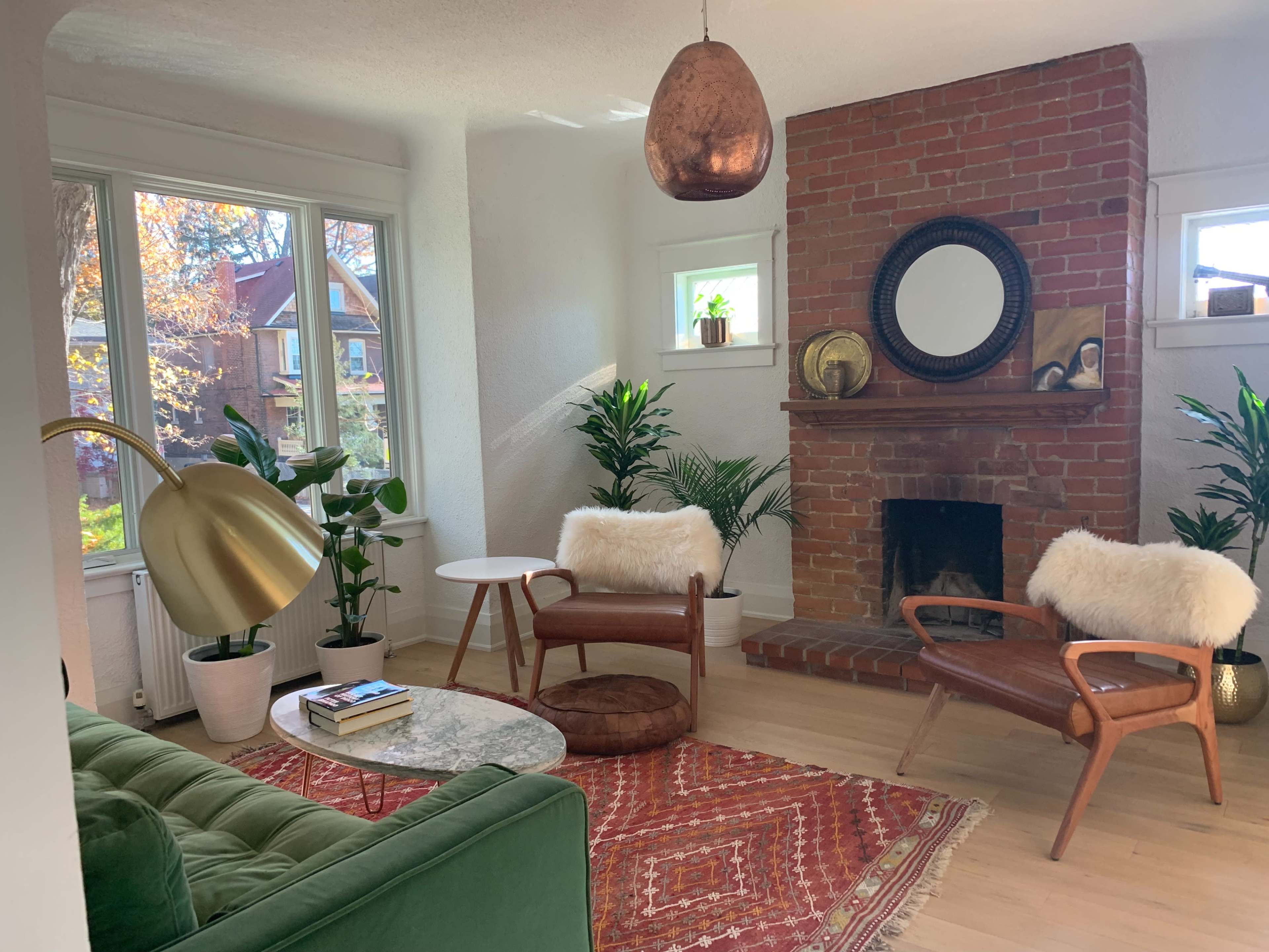 A cozy living room features a green couch, a round marble coffee table, wooden chairs with fur cushions, and large plants near a brick fireplace.