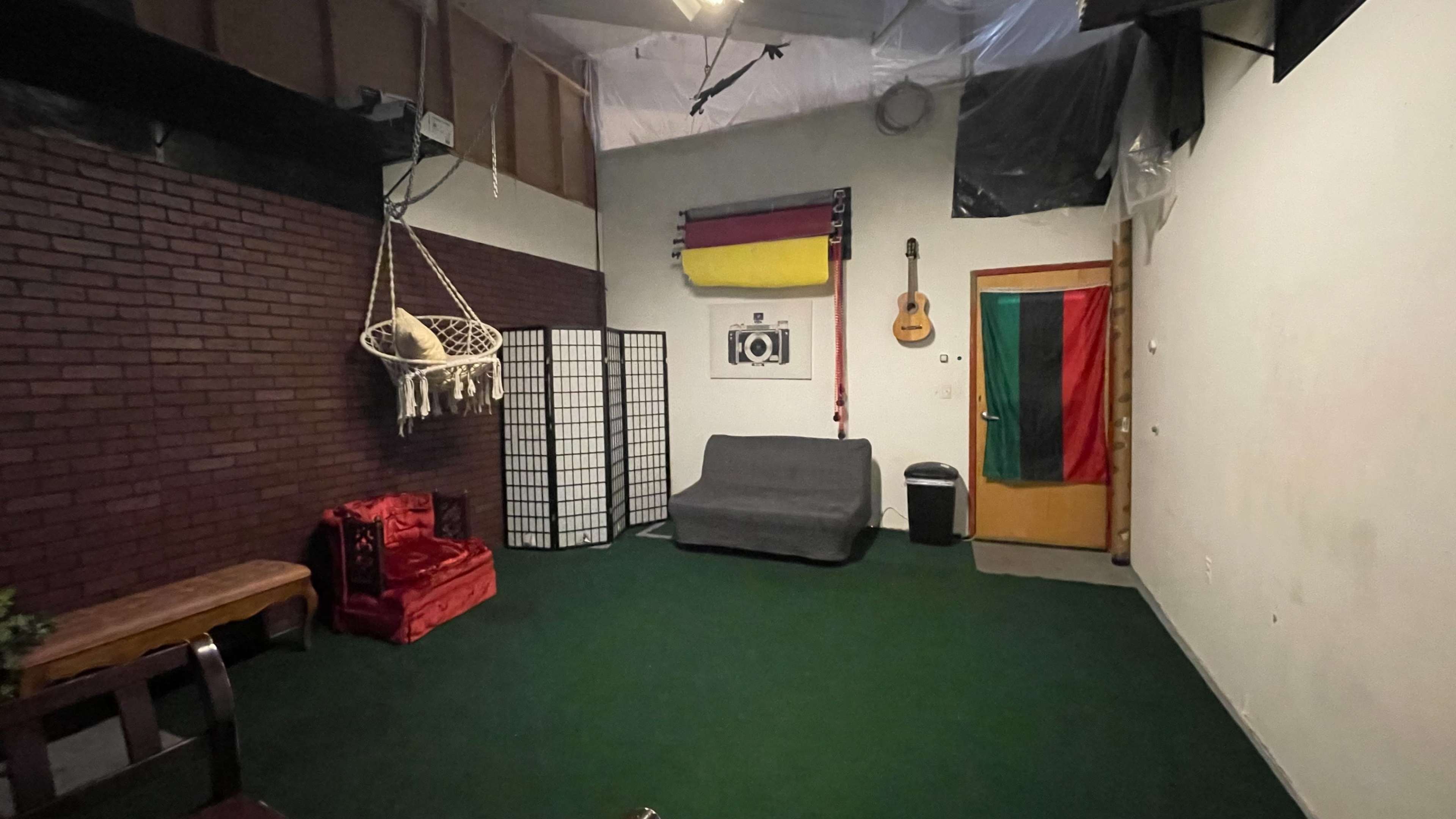 The image shows a room with green carpet, a gray couch, a hanging chair, and a door with flags and a guitar on the wall.