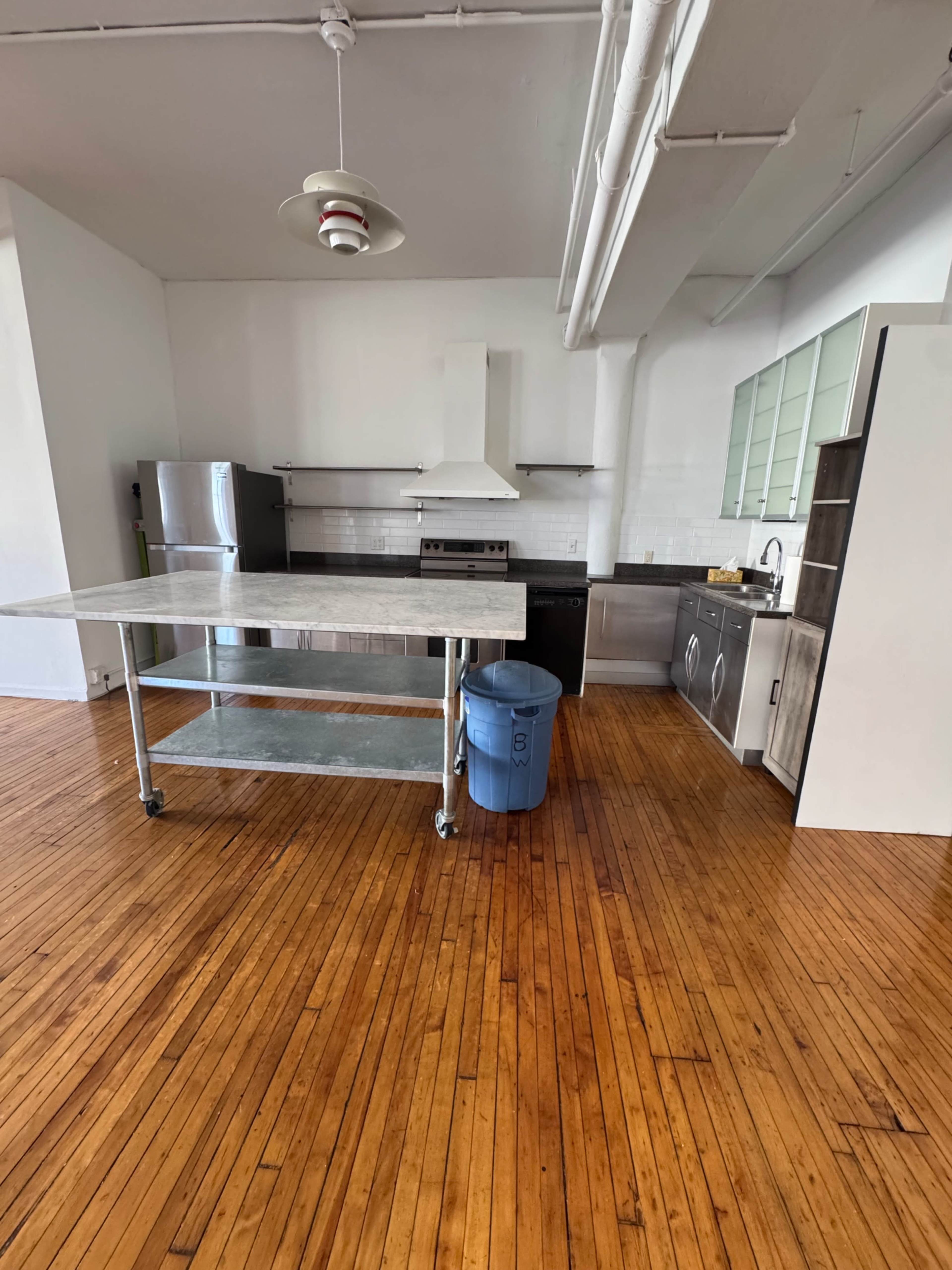 The image shows a modern kitchen with stainless steel appliances, a wooden floor, a large island on wheels, and a blue trash bin.