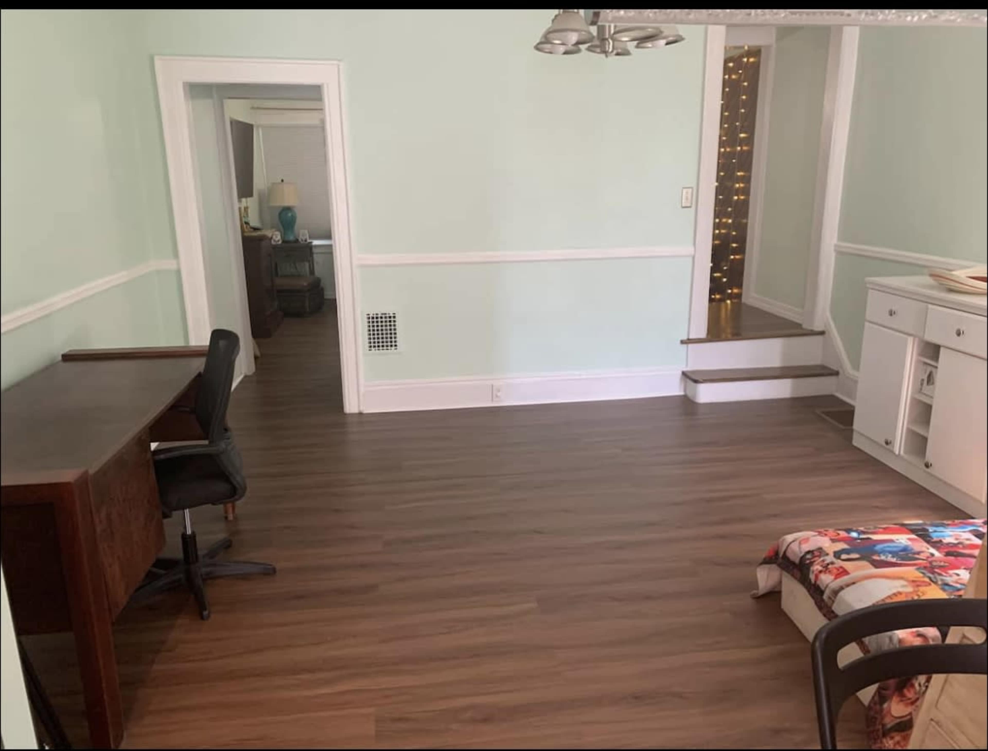 A light green room with wooden flooring, featuring a black desk and chair, a small patterned blanket on the floor, and an open doorway leading to another room.