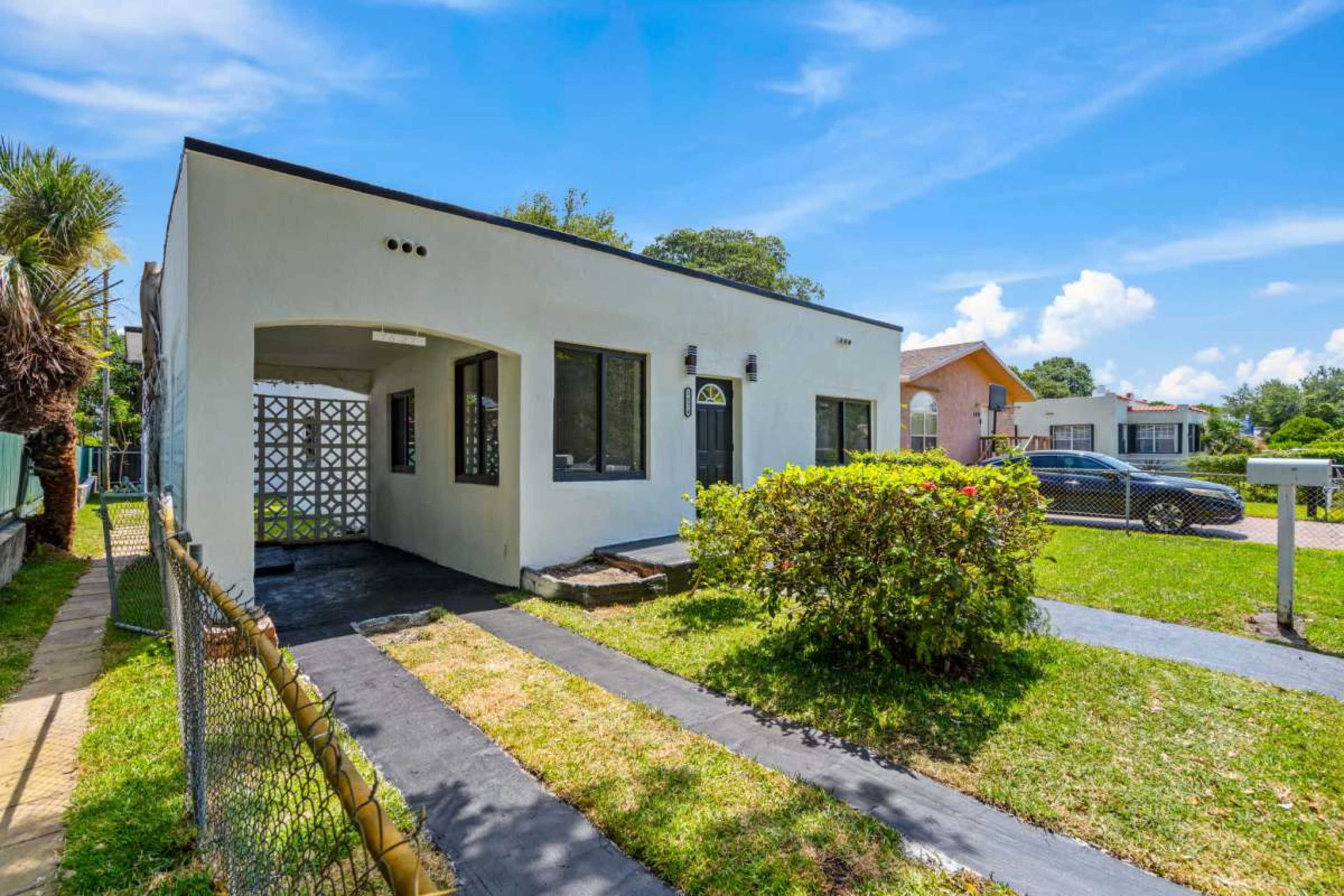The image shows a single-story house with a simple white exterior, a small front garden bordered by a fence, and a paved walkway leading to the front door.