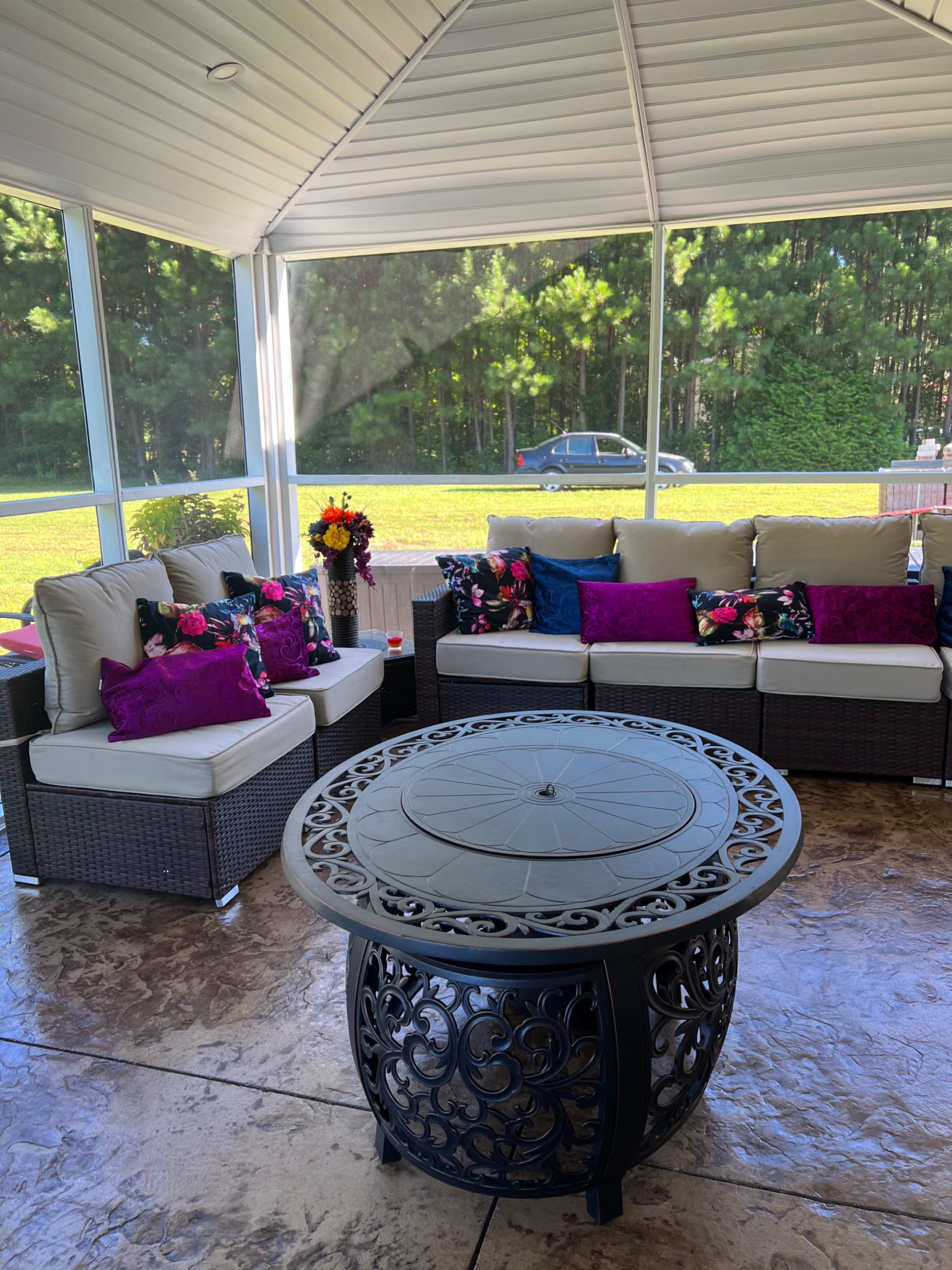 A screened-in porch features a seating area with white cushions and colorful pillows around a circular metal coffee table, surrounded by greenery.