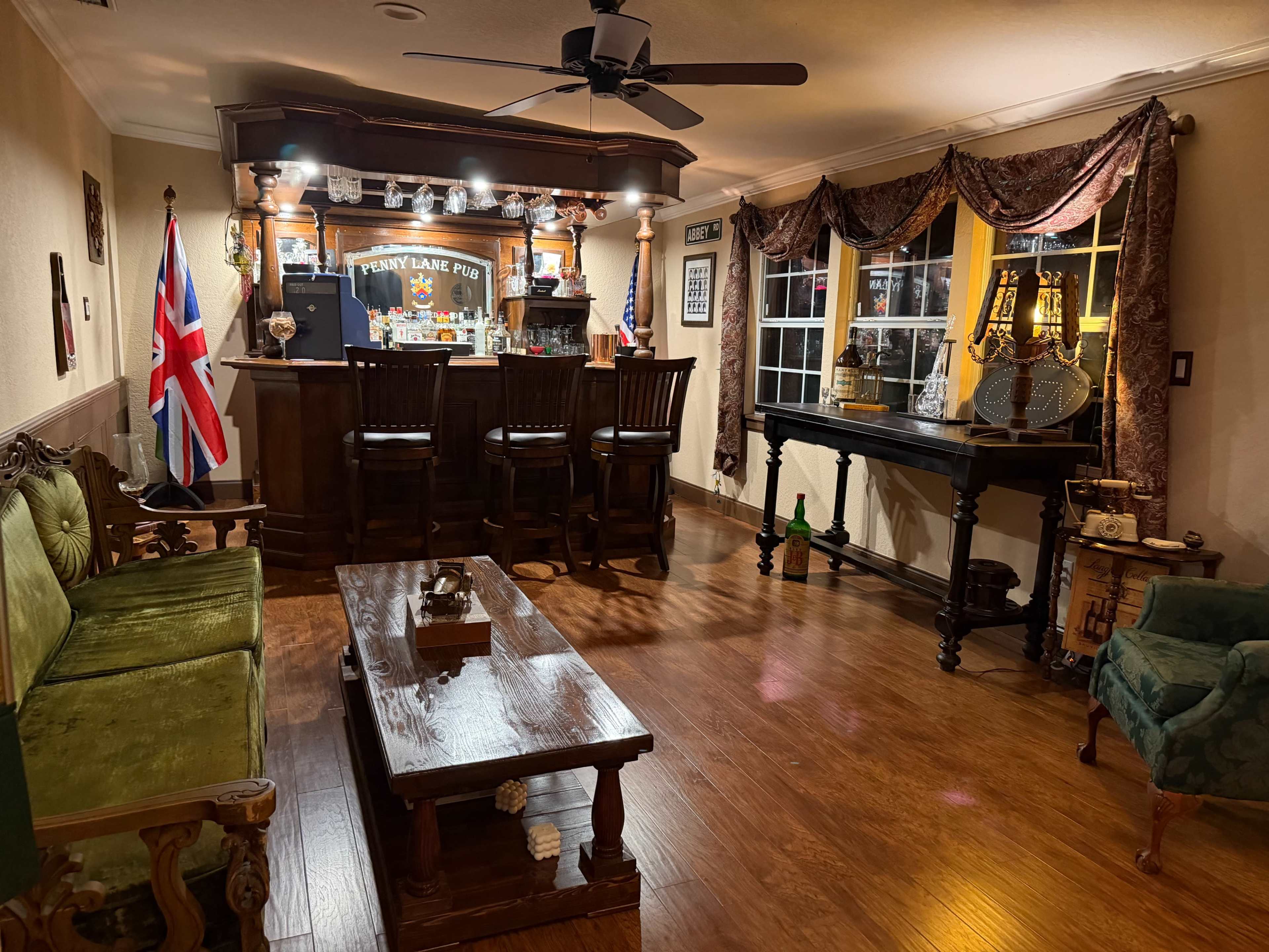 The image shows a cozy home bar area featuring a wooden bar with high stools, several flags, and decorative elements, all set in a warmly lit room with wooden flooring.