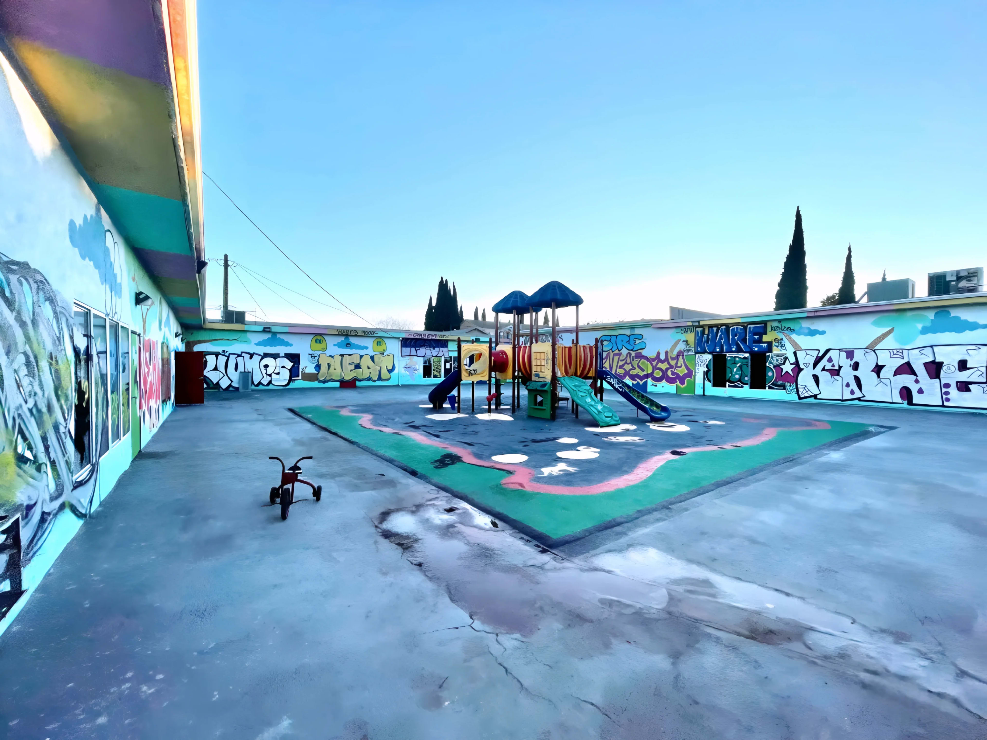 The image shows an empty playground with colorful equipment and graffiti-covered walls under a clear blue sky.