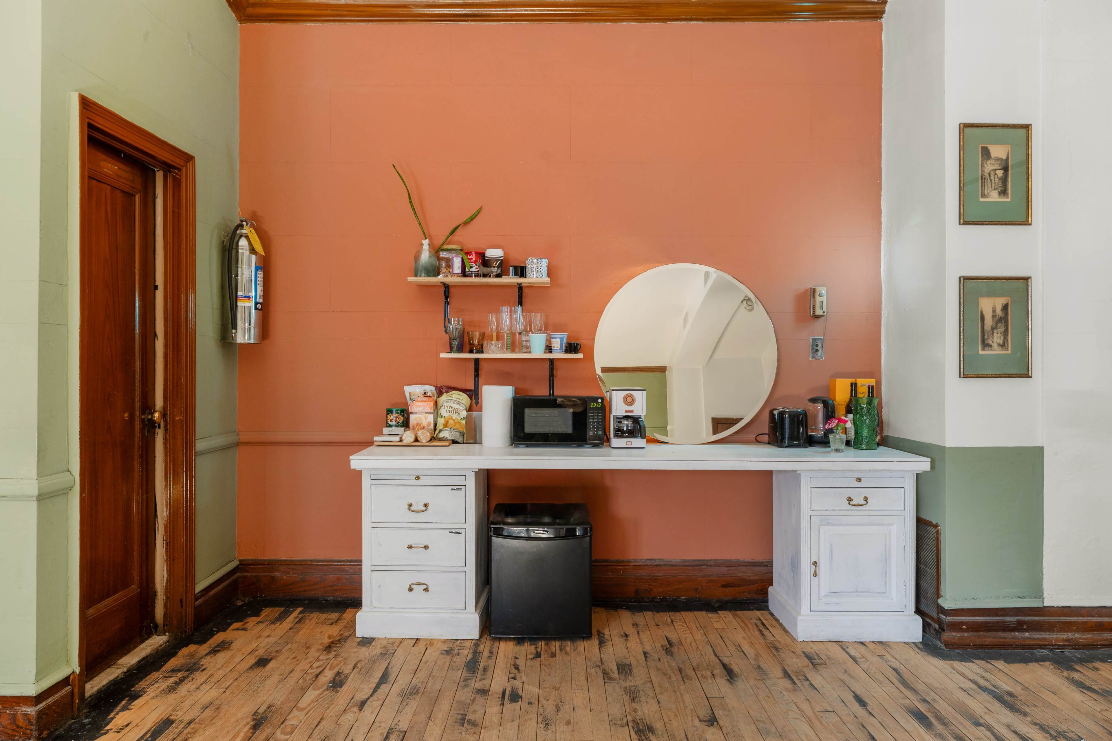 The image shows a small kitchenette area with a white counter, a mirror, shelves containing various items, and a microwave, situated against a peach-colored wall.