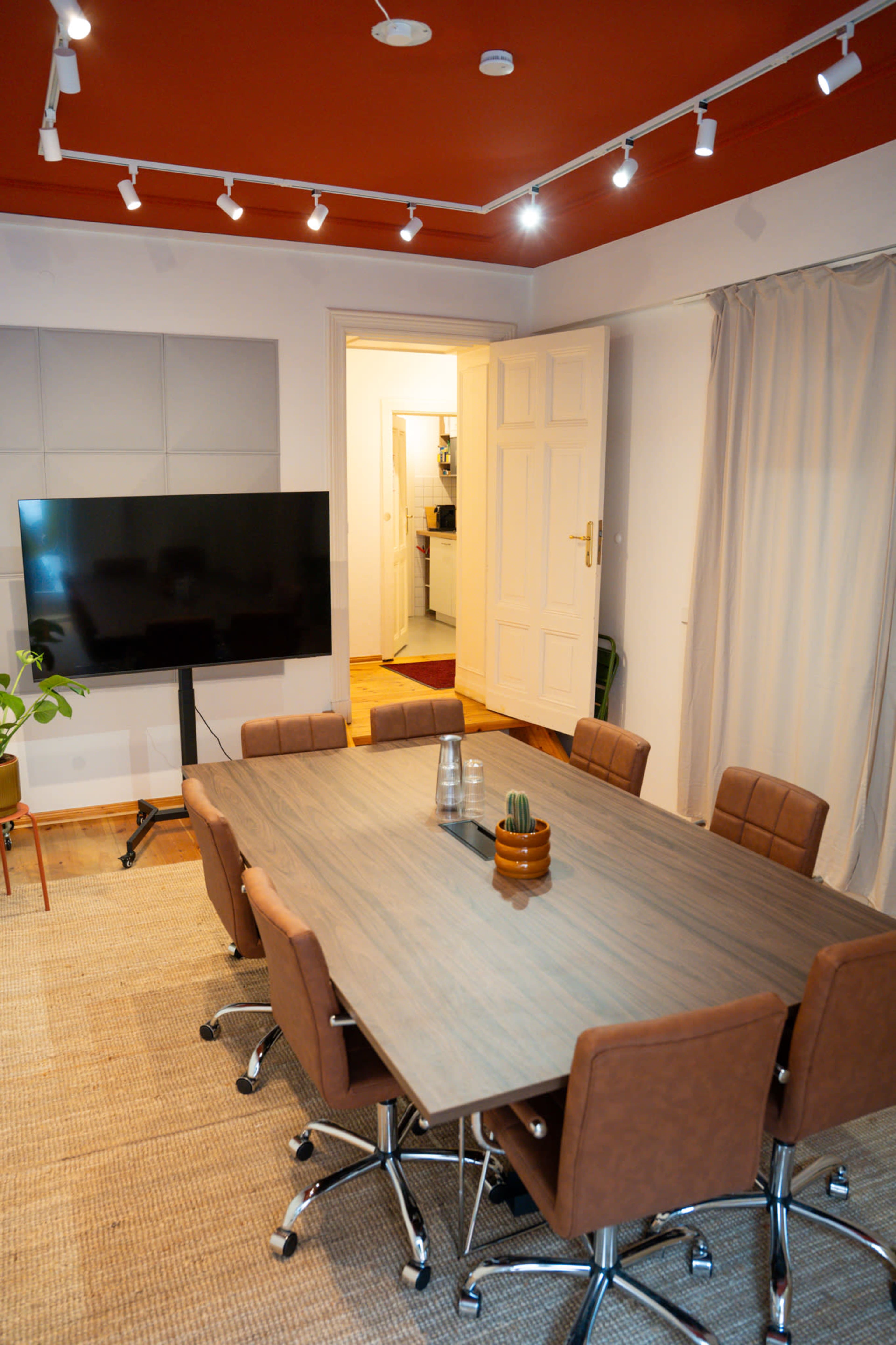 A modern conference room features a large wooden table surrounded by brown leather chairs, with a television mounted on a stand and a doorway leading to another room.