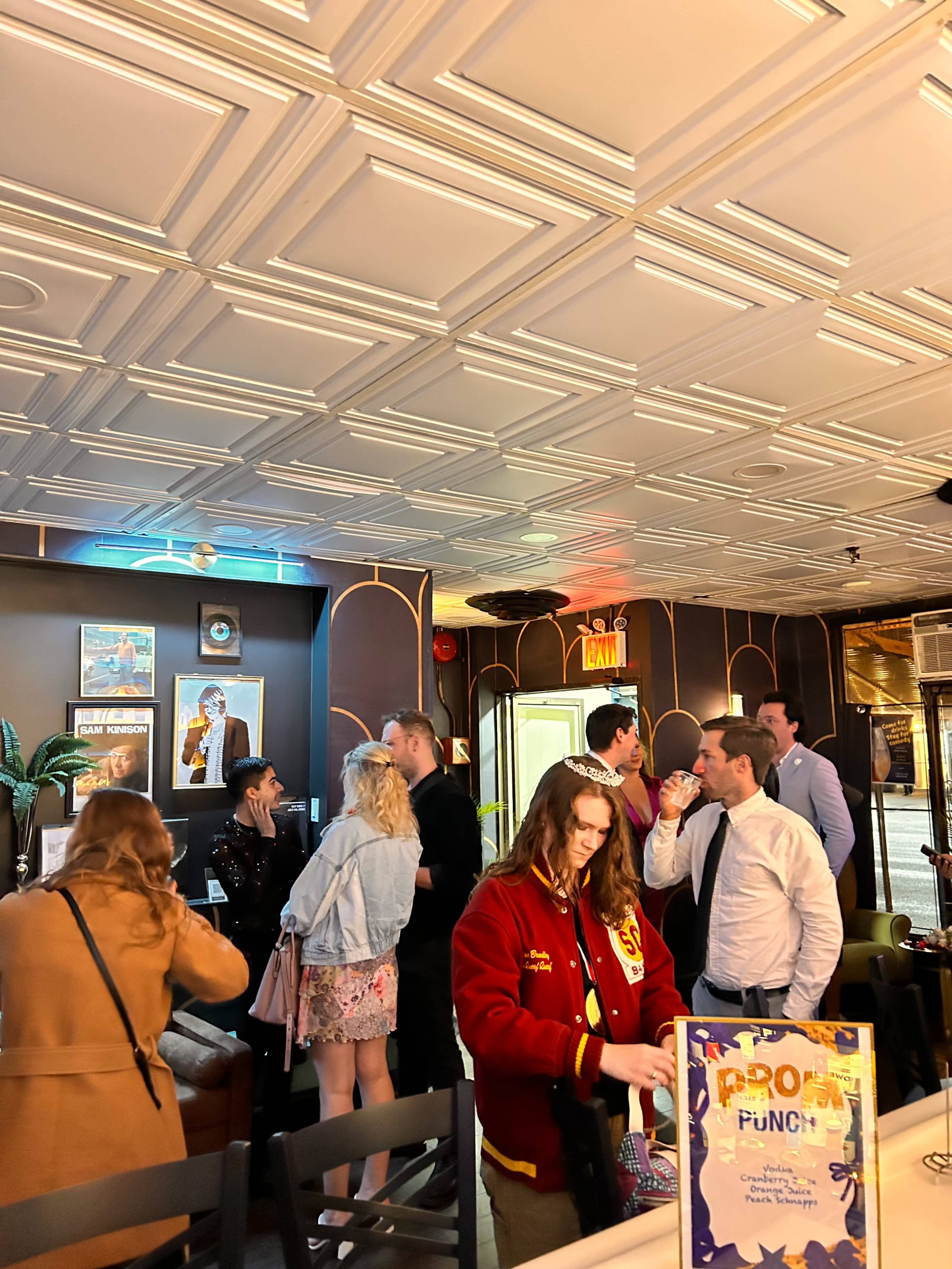 A group of people stands in a lively bar setting with decorative ceilings and a mix of casual and formal attire.