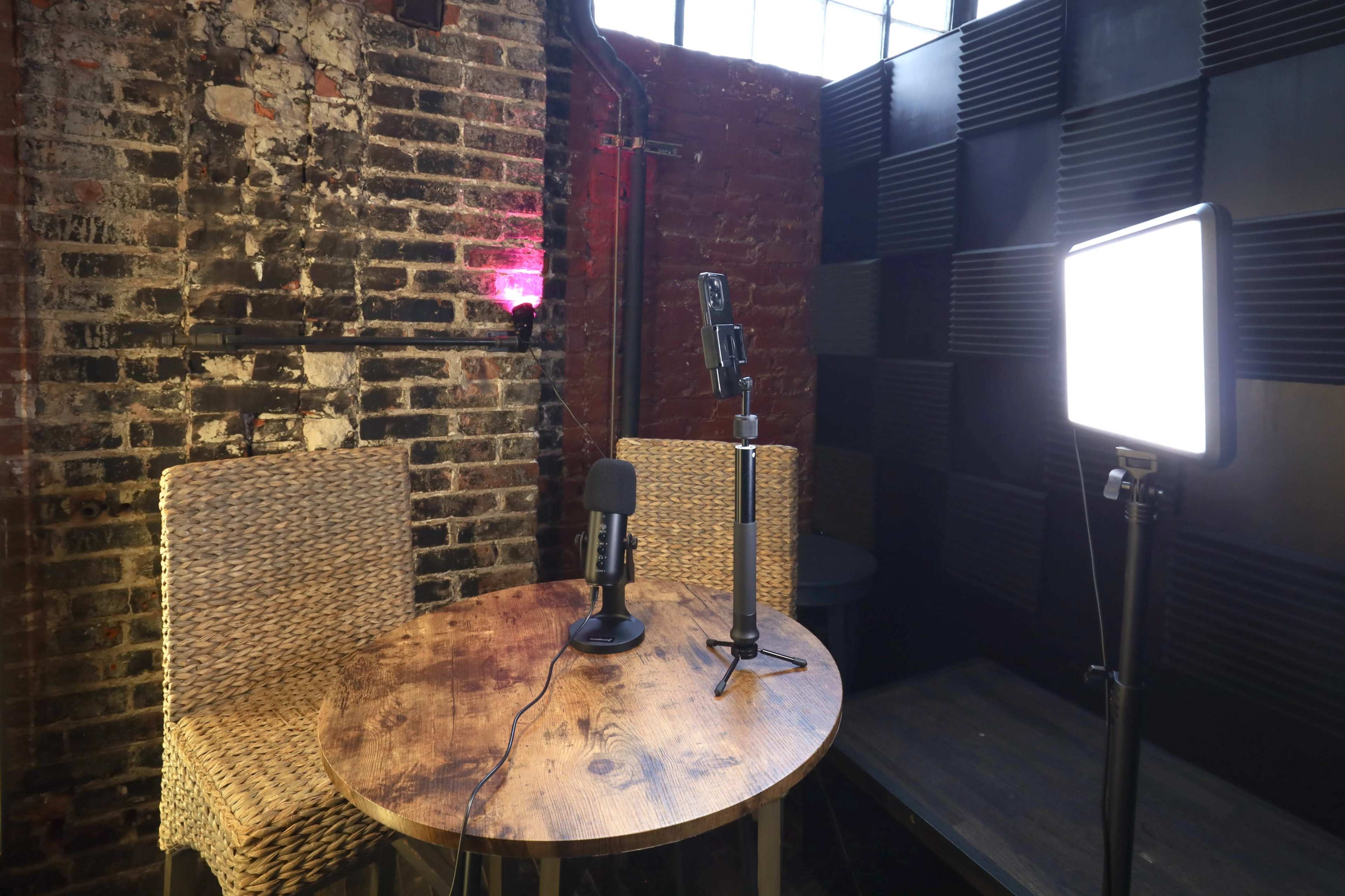 The image shows a small recording setup with a round wooden table, two wicker chairs, a microphone on a stand, and a bright lighting panel against a textured brick wall.