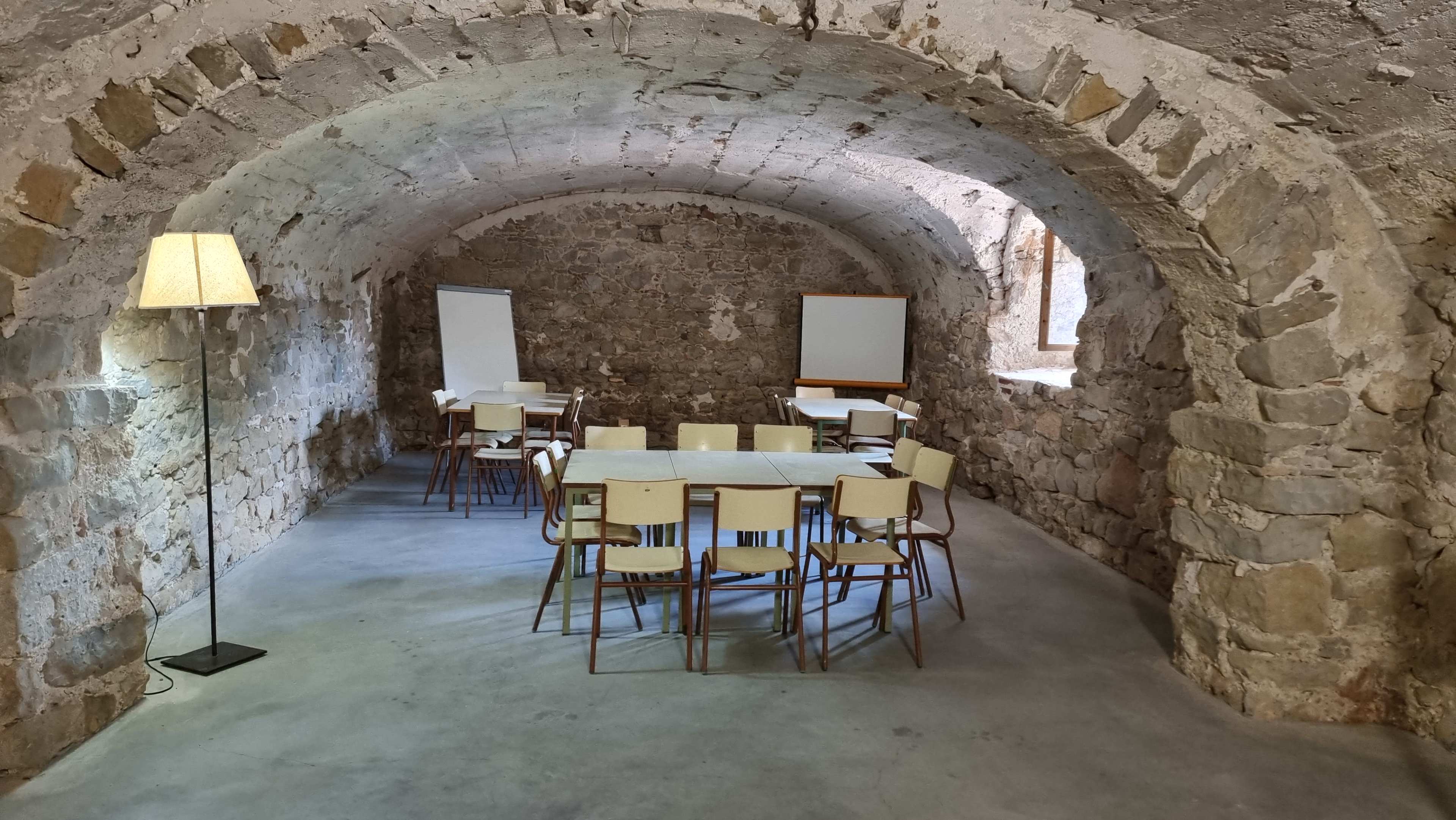 The image shows a stone-walled seminar room with a long table, chairs arranged around it, and two flip charts on easels at one end.