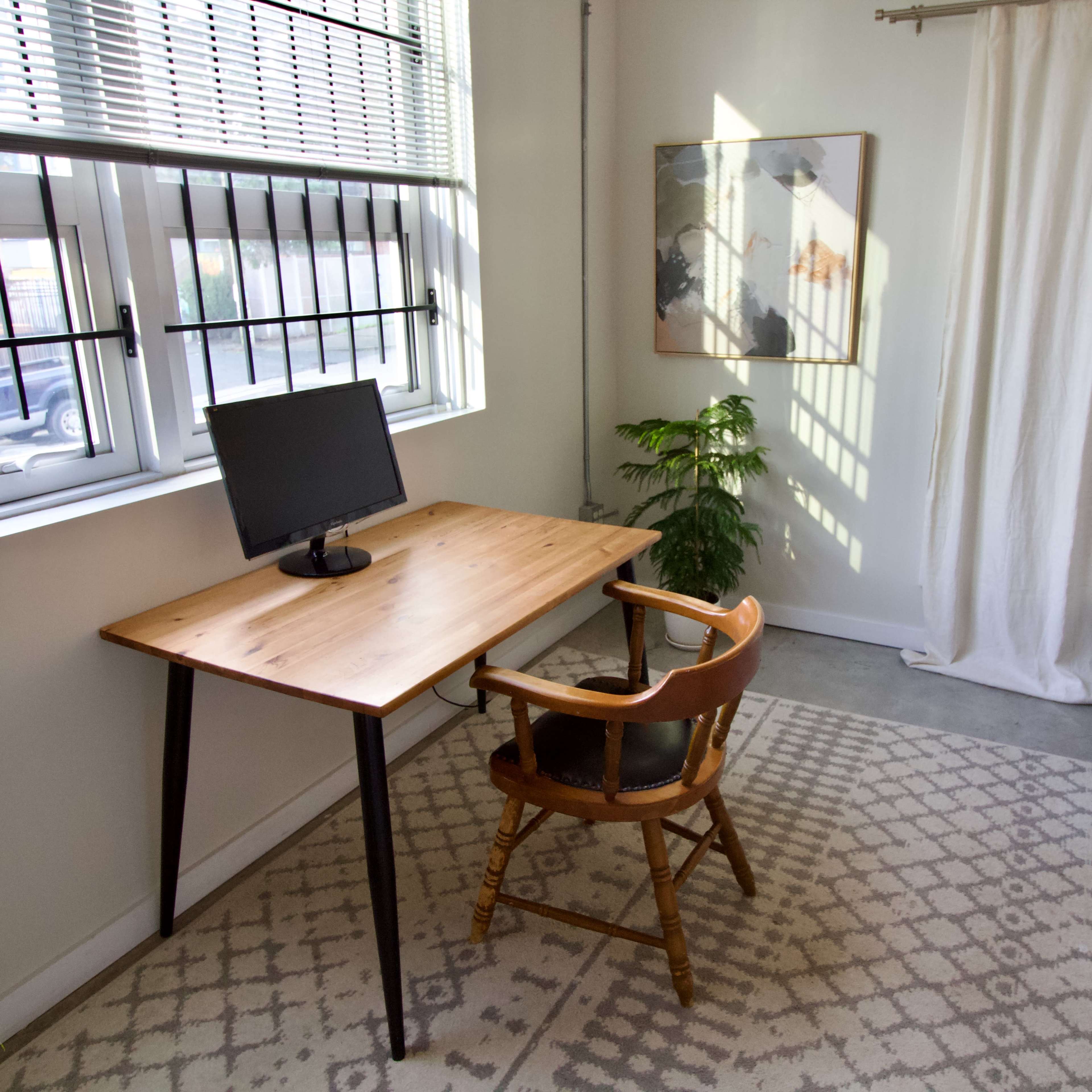 A wooden desk with a computer monitor and a wooden chair is positioned near a window, next to a potted plant in a bright room.