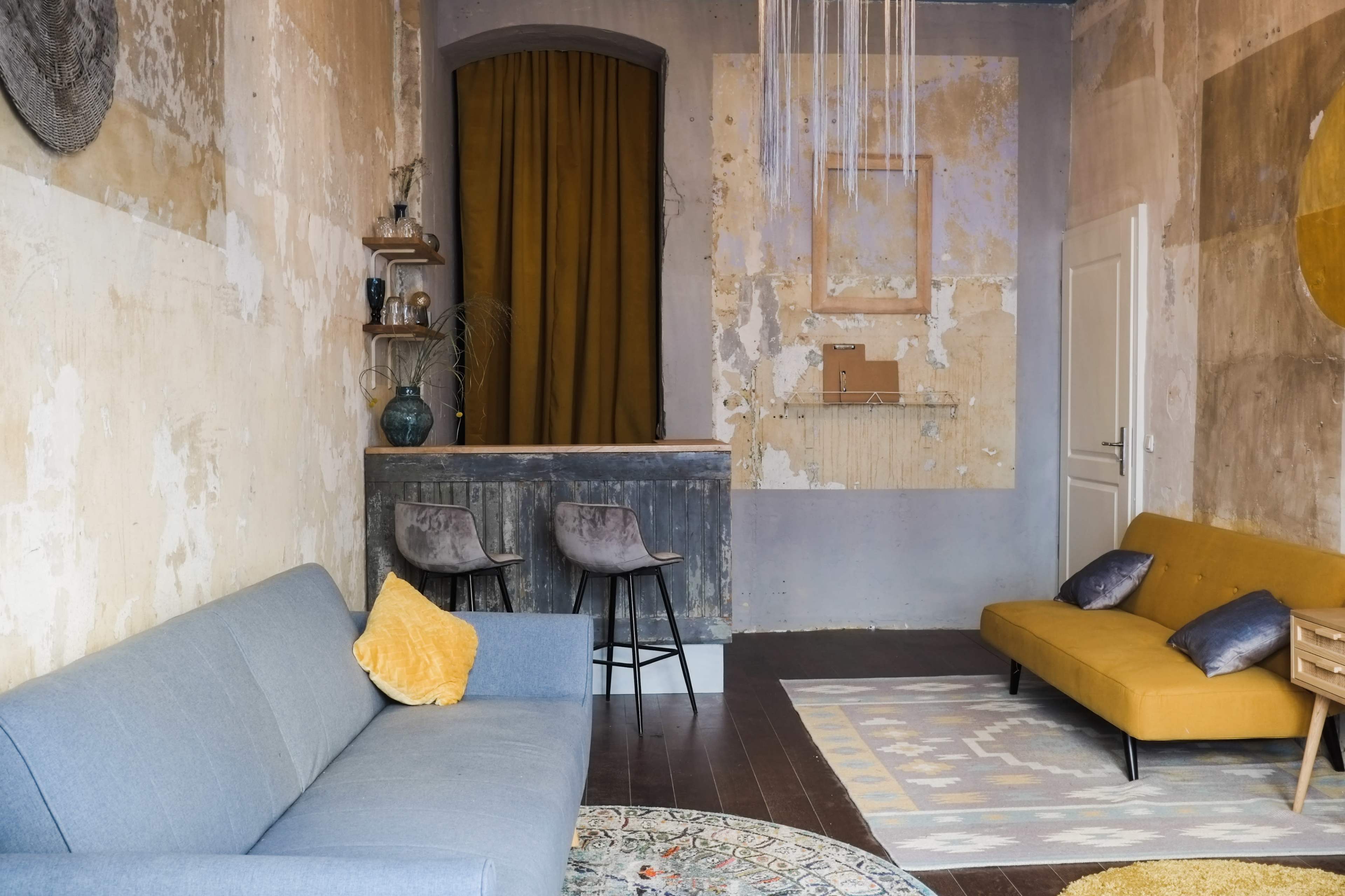 A room with a gray sofa, a yellow seating area, a small bar with two stools, and walls featuring peeling paint and a large circular decoration.