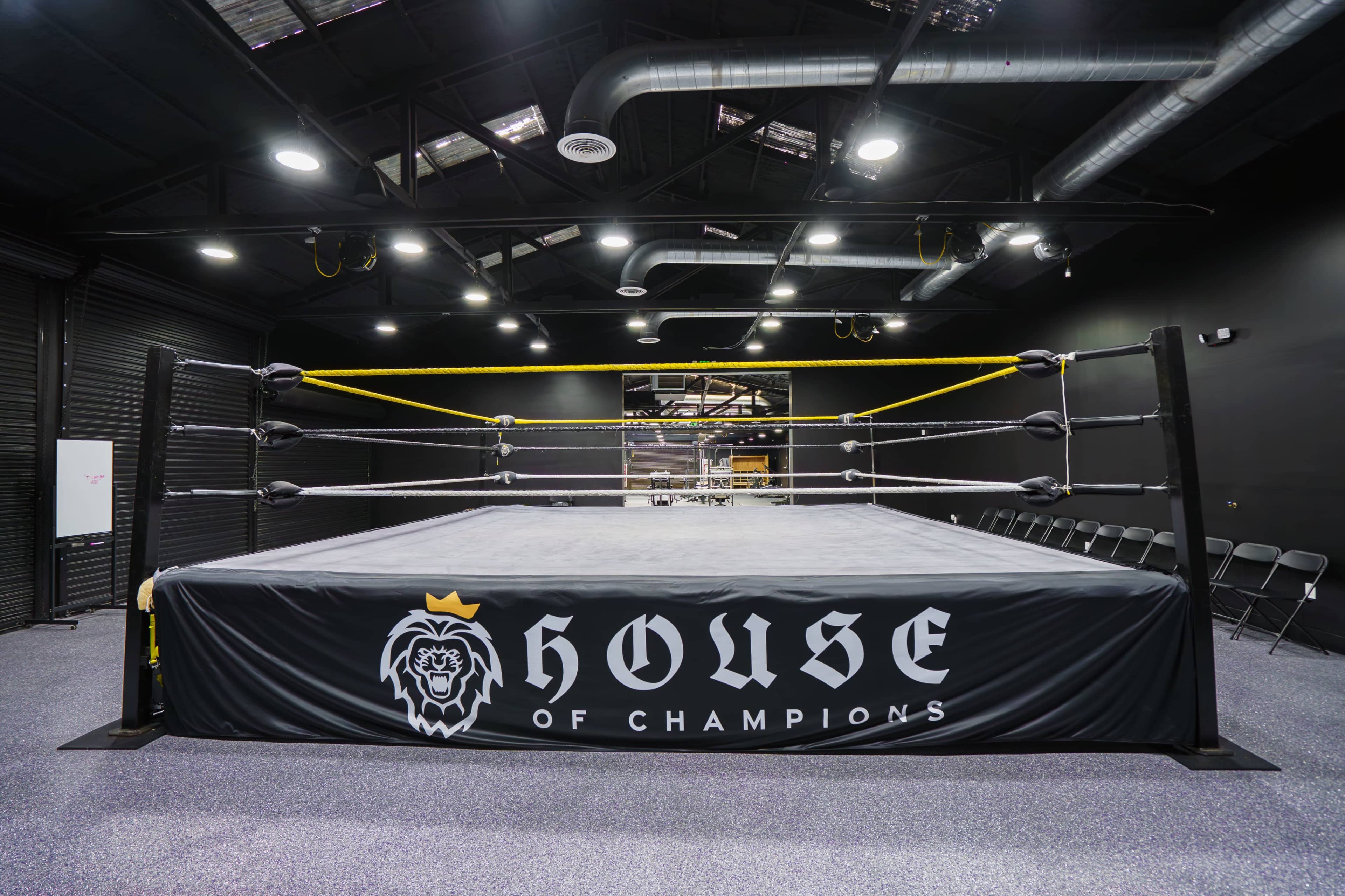 The image shows a wrestling ring inside a dark, modern training facility with black walls and a logo for "House of Champions" on the ring mat.