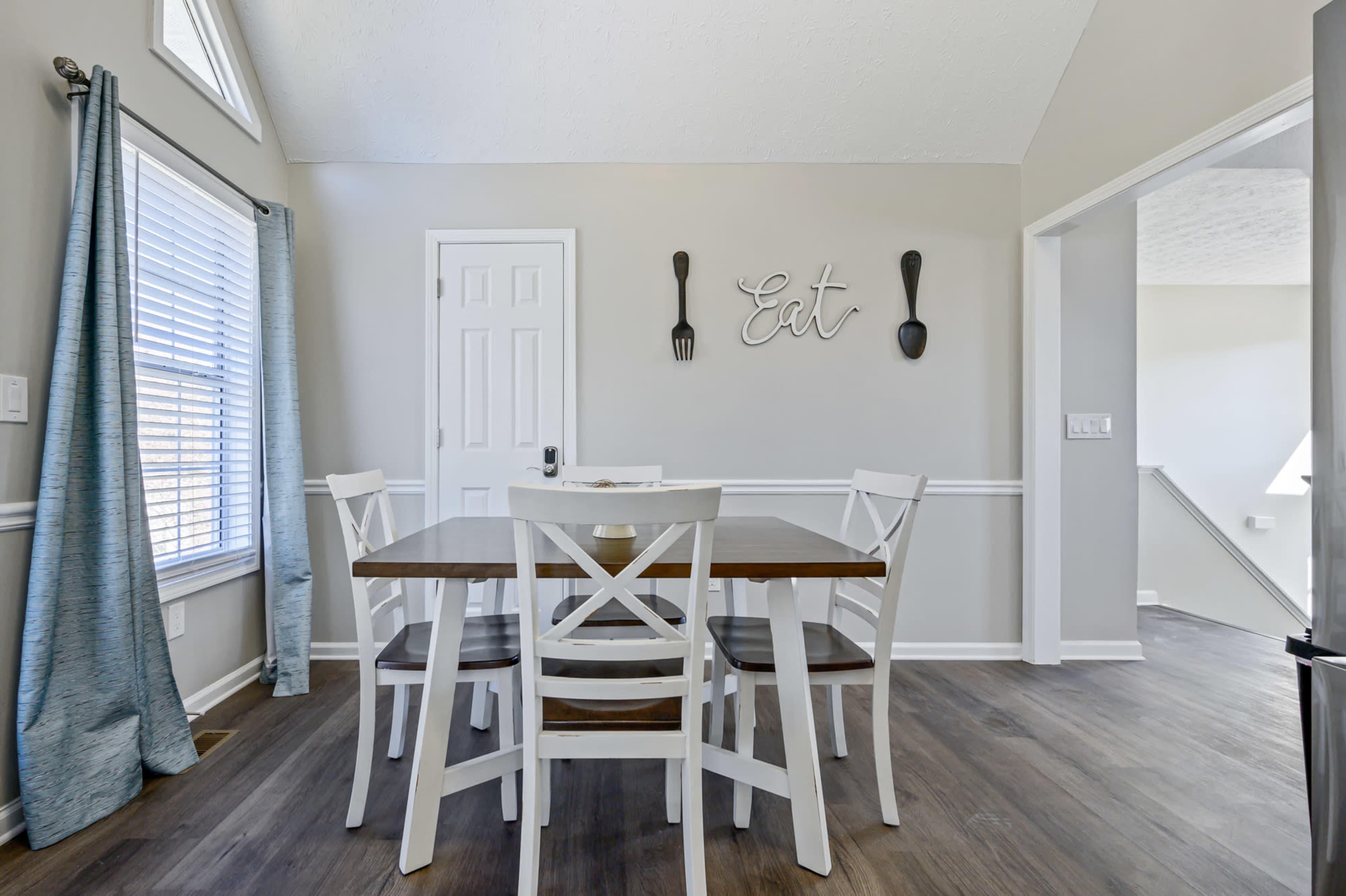 A dining area features a wooden table surrounded by white chairs, with a wall decoration that says "Eat" and a door leading to another space.