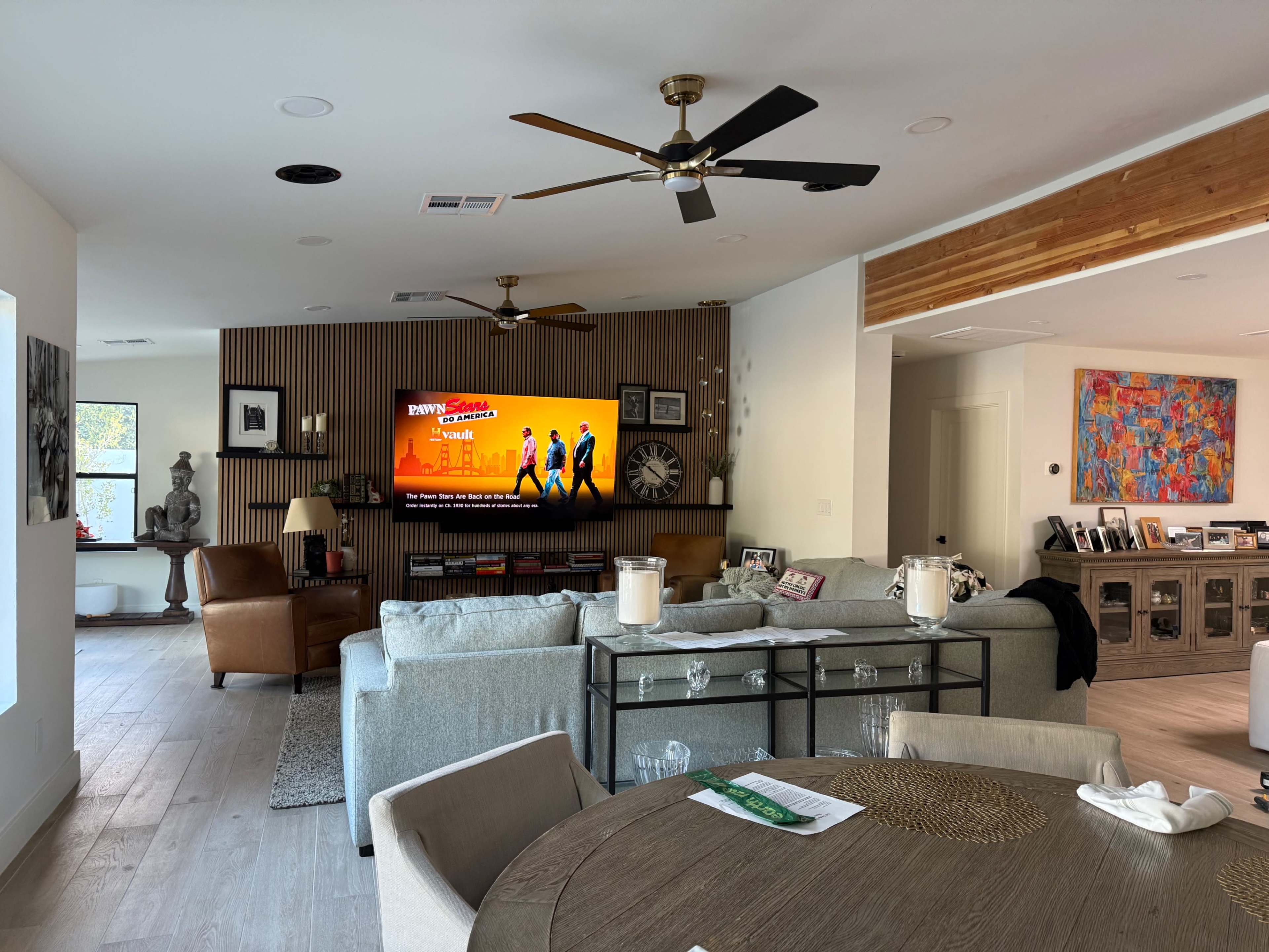 The image shows a modern living room with a large television mounted on the wall, a sofa, a dining table, and various decor elements.
