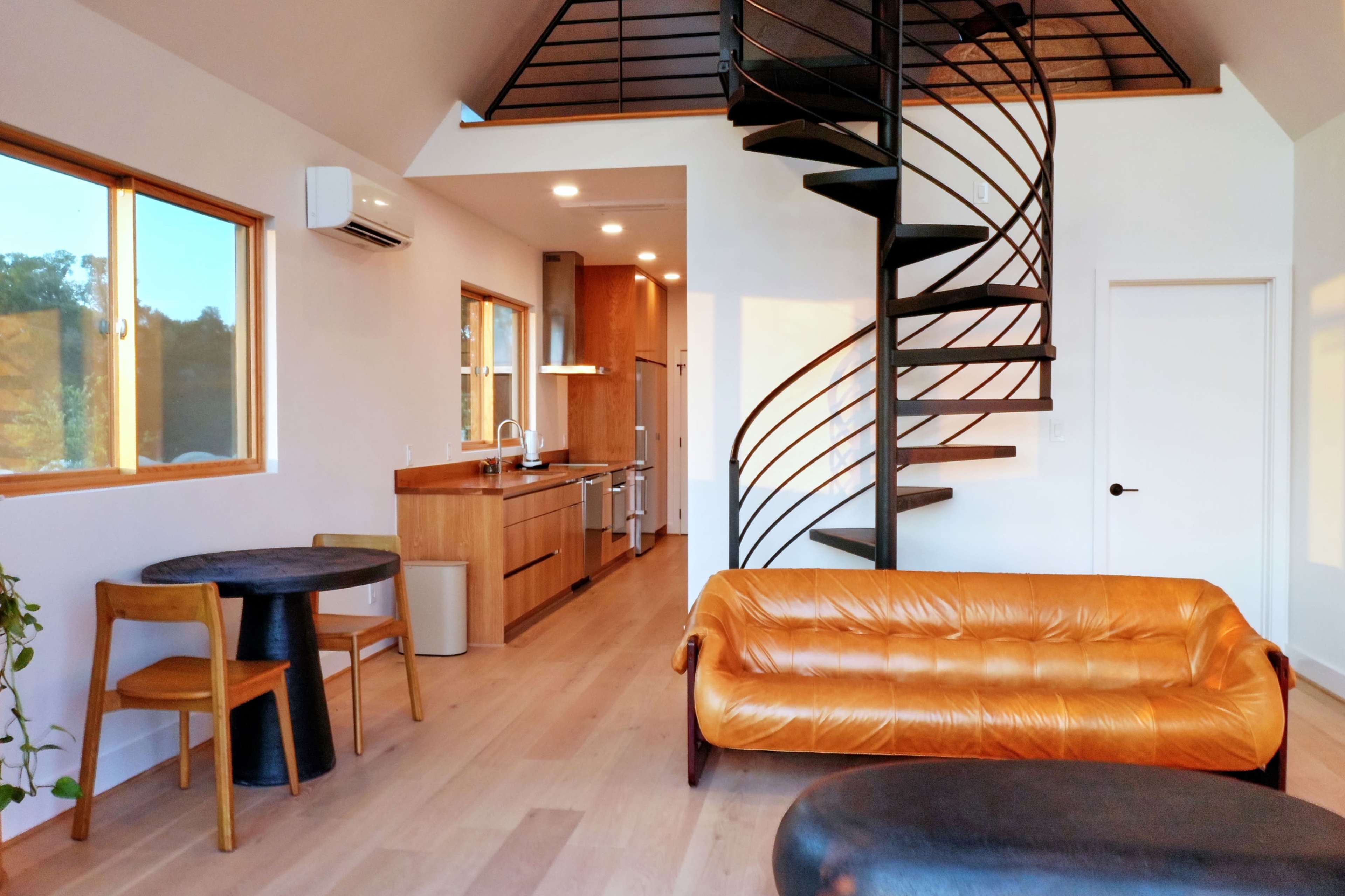 The image shows a modern living space featuring a spiral staircase, a beige leather couch, a round black table with two chairs, and a kitchen area in the background.