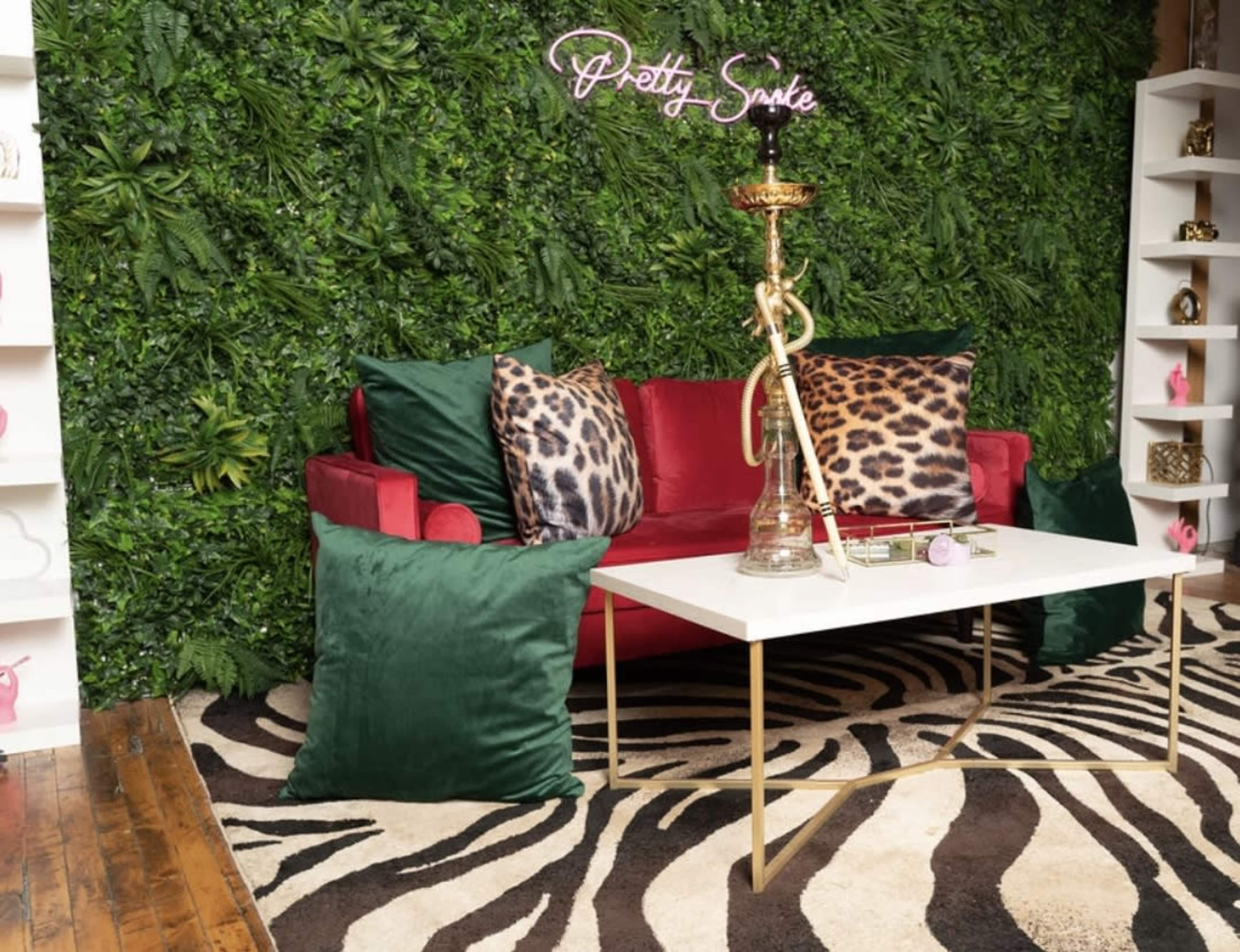 A red couch with leopard print pillows is positioned in front of a green wall with a neon sign, alongside a coffee table and a hookah on the table.