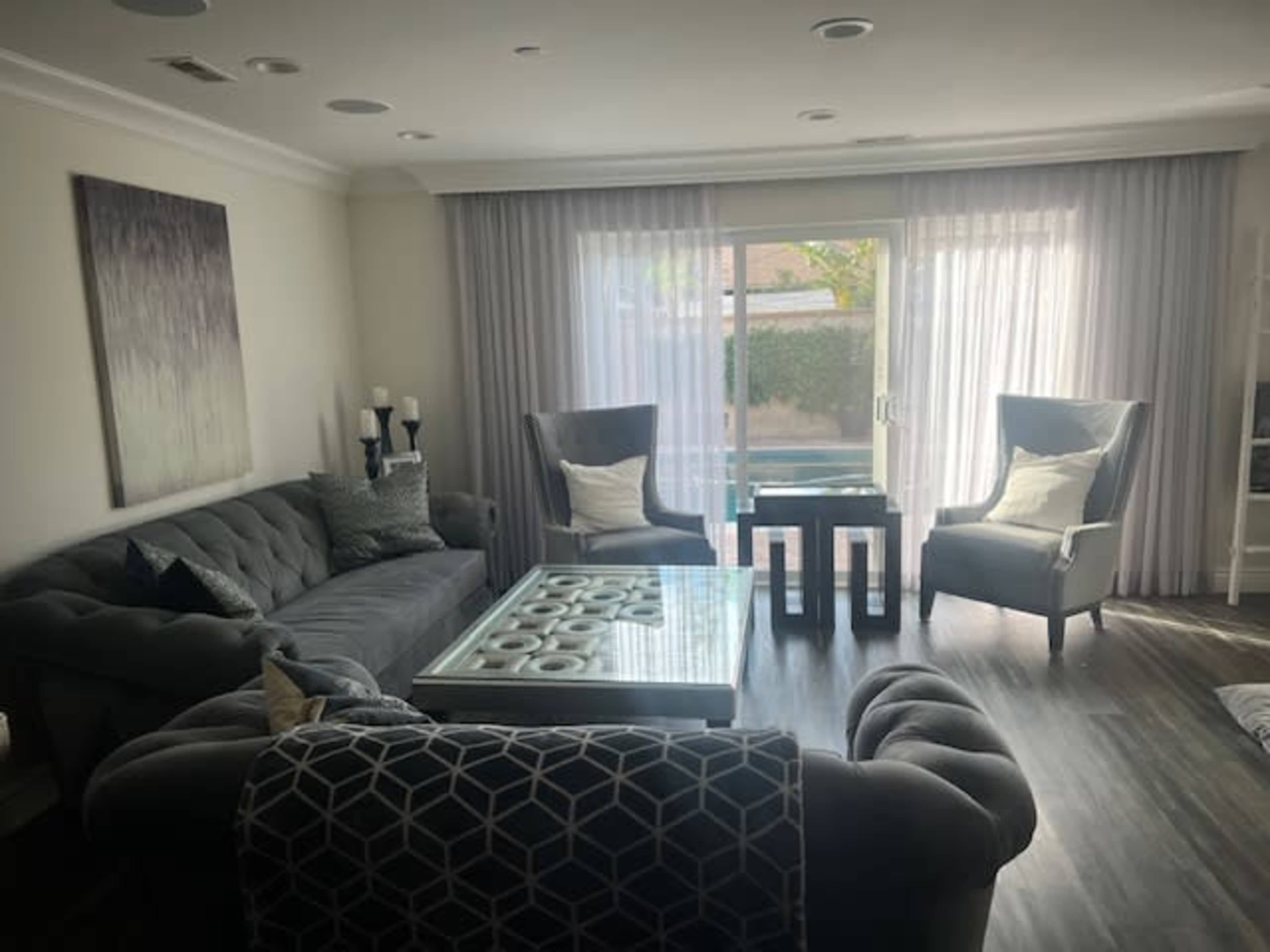 A modern living room features a plush gray sofa, two large armchairs, a glass coffee table, and large windows with sheer curtains allowing natural light.