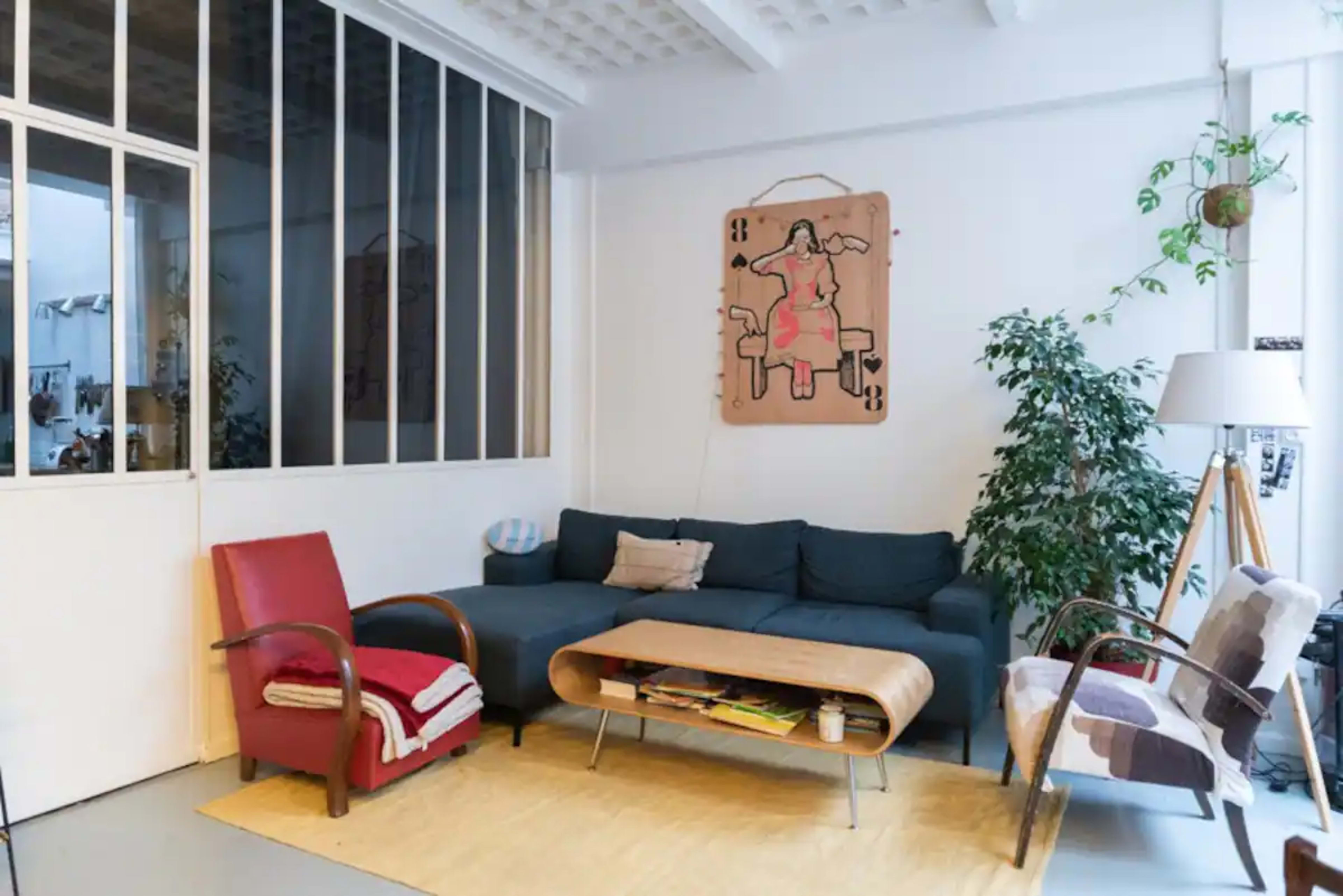 The image shows a modern living room with a blue sofa, a wooden coffee table, and a red armchair, surrounded by plants and a large window.
