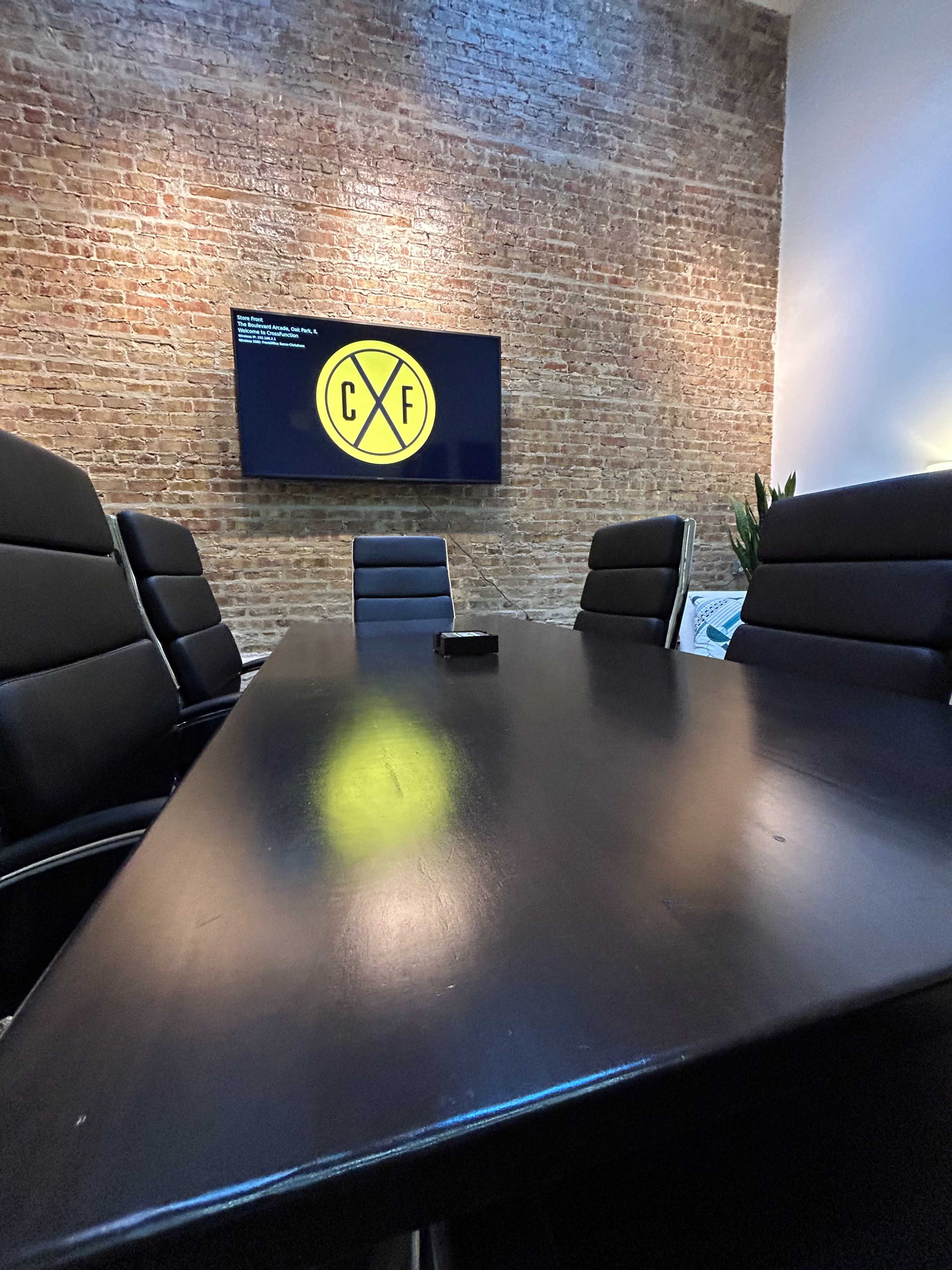 A conference room features a long black table surrounded by chairs, with a TV mounted on a brick wall displaying a yellow logo.